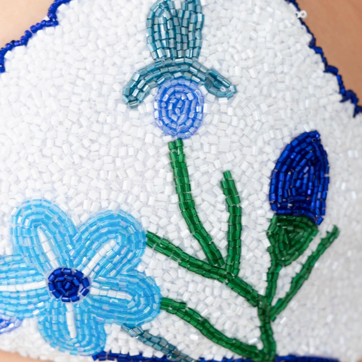 Close-up of the SELF-ADORE Euro Summer Hand-Beaded Embellished Bikini Top in blue, featuring intricate blue flower and green stem beadwork on white—perfect for chic European summer swimwear.