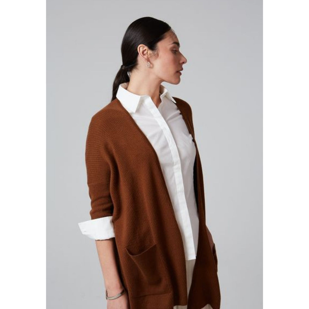 A woman with dark hair tied back wears a white shirt and the Syrup Brown Lillien Open Cardigan by OO-TO, handcrafted in Nepal, standing against a plain gray background and looking to the side.