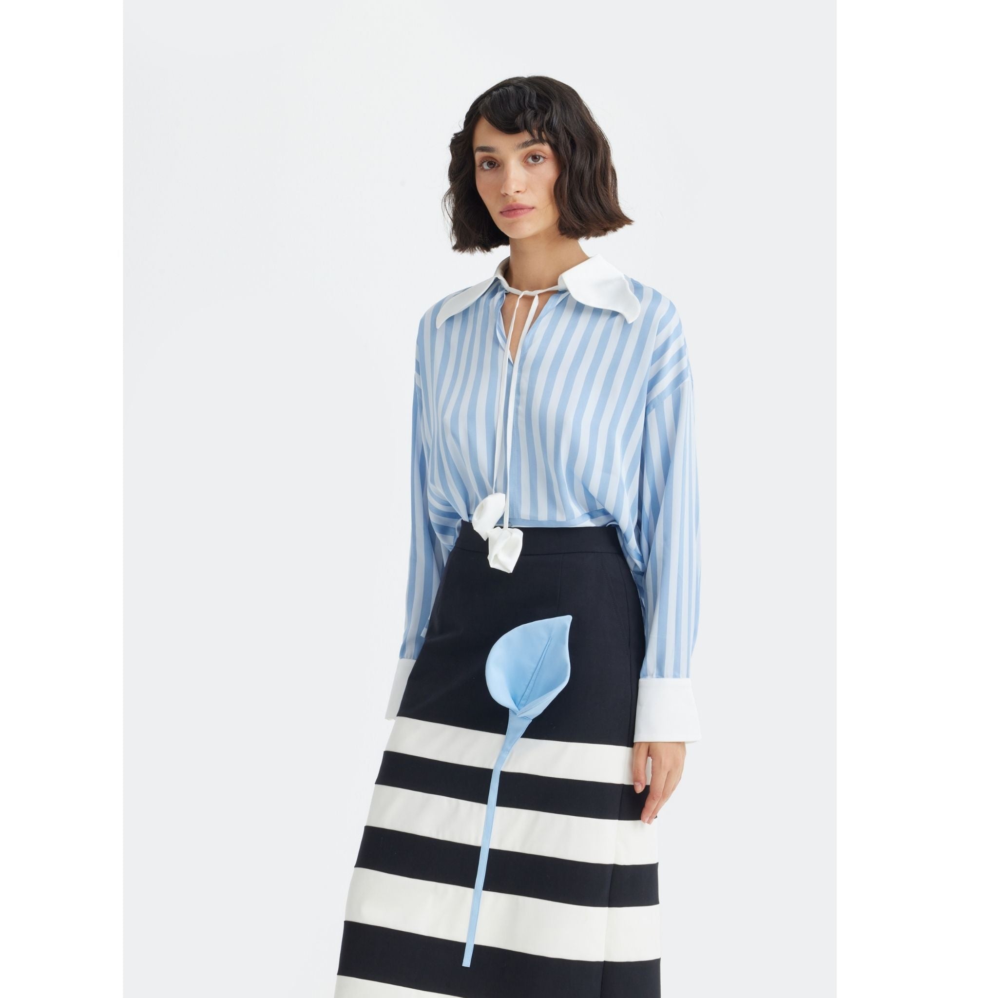 A woman with short dark hair wears the JULIA ALLERT Smart Elegance – Silk-Touch Striped Long-Sleeve Blouse in Light Blue, featuring an oversized white collar and cuffs, paired with a black and white striped skirt against a plain light background.
