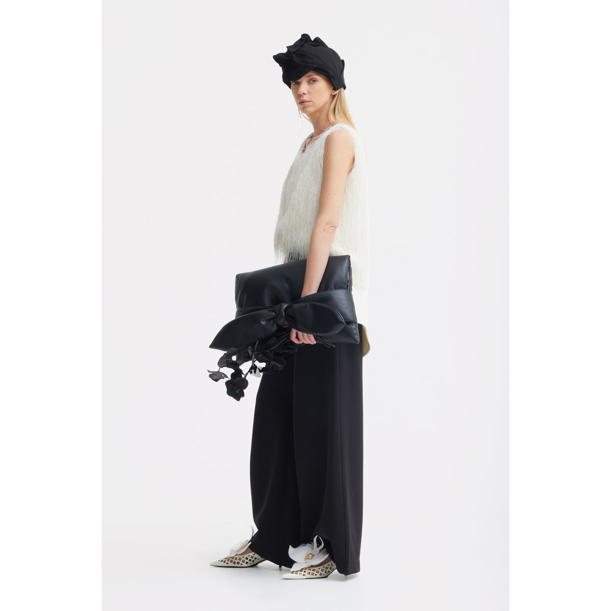 A woman in a white fringed sleeveless top, JULIA ALLERT Statement Waist Wide-Leg Trousers in black, white pointed shoes, and a black headwrap holds a large black clutch with bow and floral accents against a plain white background.