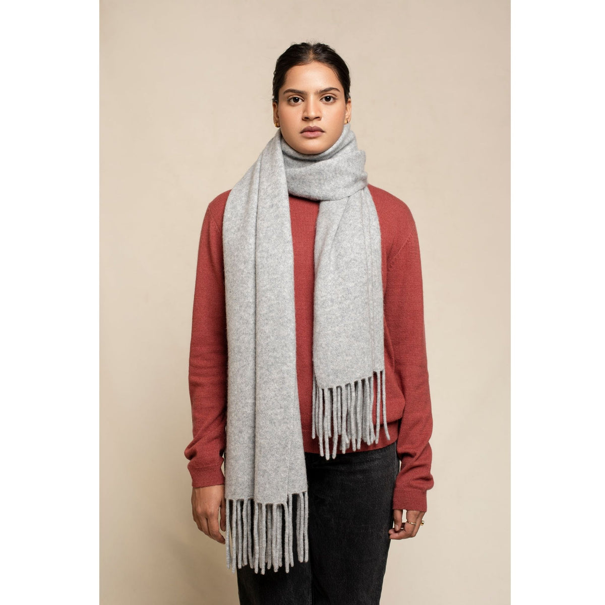 A person stands against a plain beige background, wearing a red sweater, black pants, and the OO-TO Grey Knitted Felted Cashmere Chunky Scarf handcrafted in Nepal with long fringes hanging down the front.