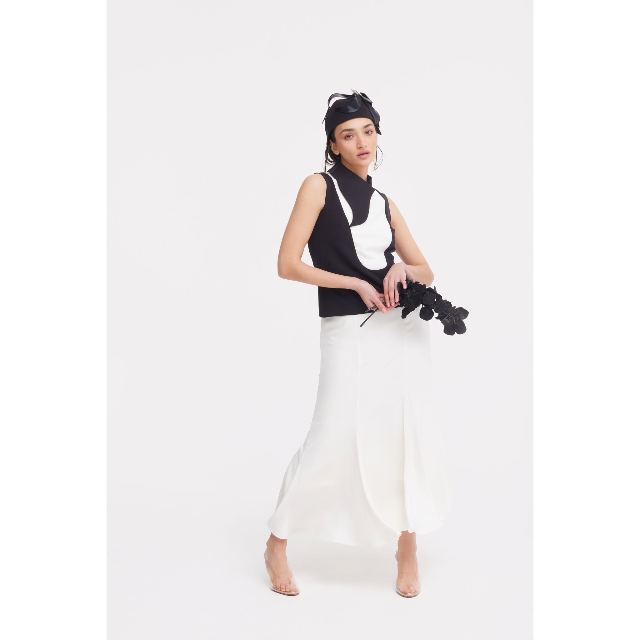 A woman in a JULIA ALLERT black and white sleeveless top, High Waist Double-Sided Satin Maxi Skirt - Ivory, and black headpiece holds black flowers against a plain white background, creating an elegant eveningwear look.