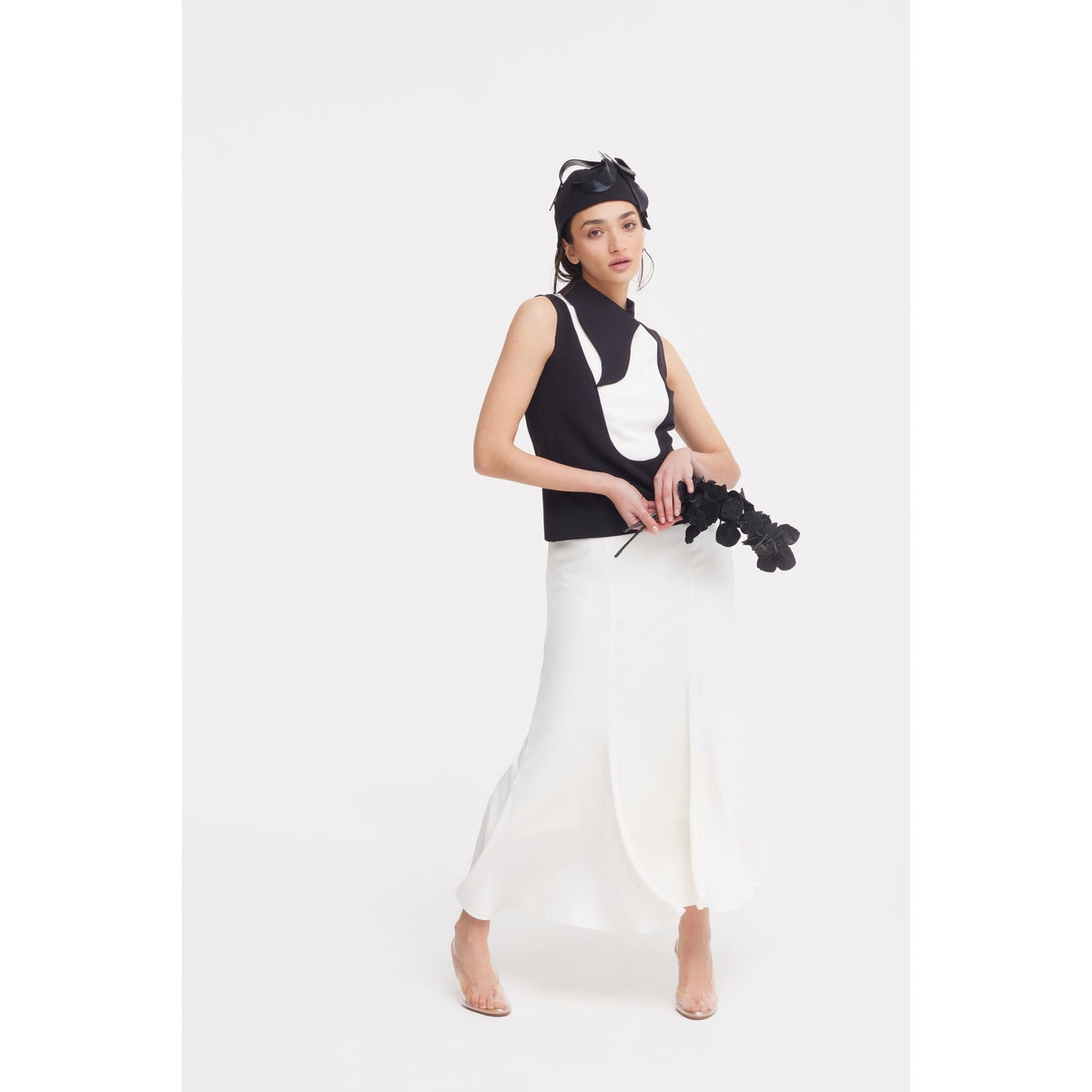 A woman in a JULIA ALLERT black and white sleeveless top, High Waist Double-Sided Satin Maxi Skirt - Ivory, and black headpiece holds black flowers against a plain white background, creating an elegant eveningwear look.