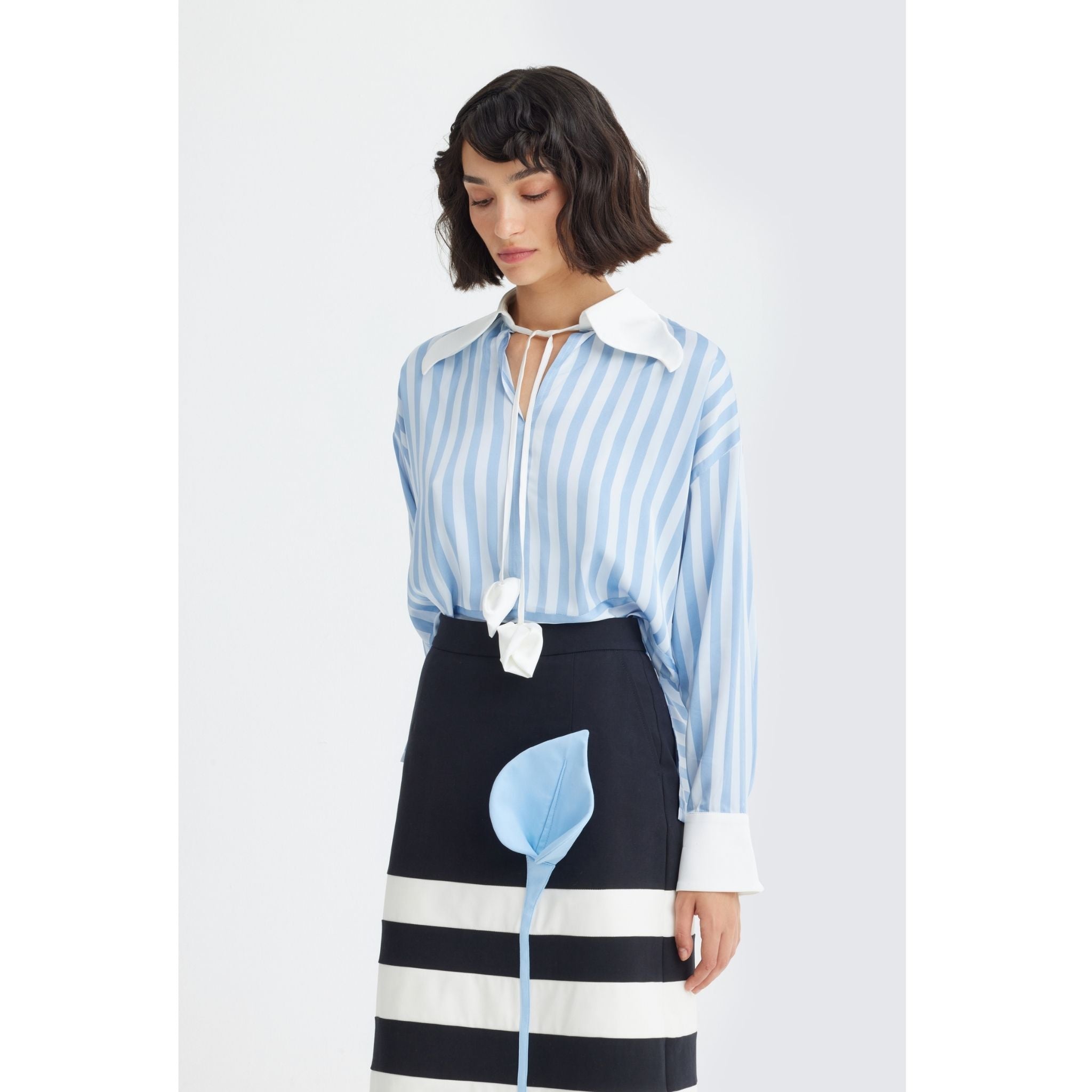 A woman with short dark hair wears the JULIA ALLERT Smart Elegance Silk-Touch Striped Long-Sleeve Blouse in light blue, featuring an oversized white collar and cuffs. She pairs it with a black skirt with wide white stripes and a blue leaf detail, looking down neutrally.