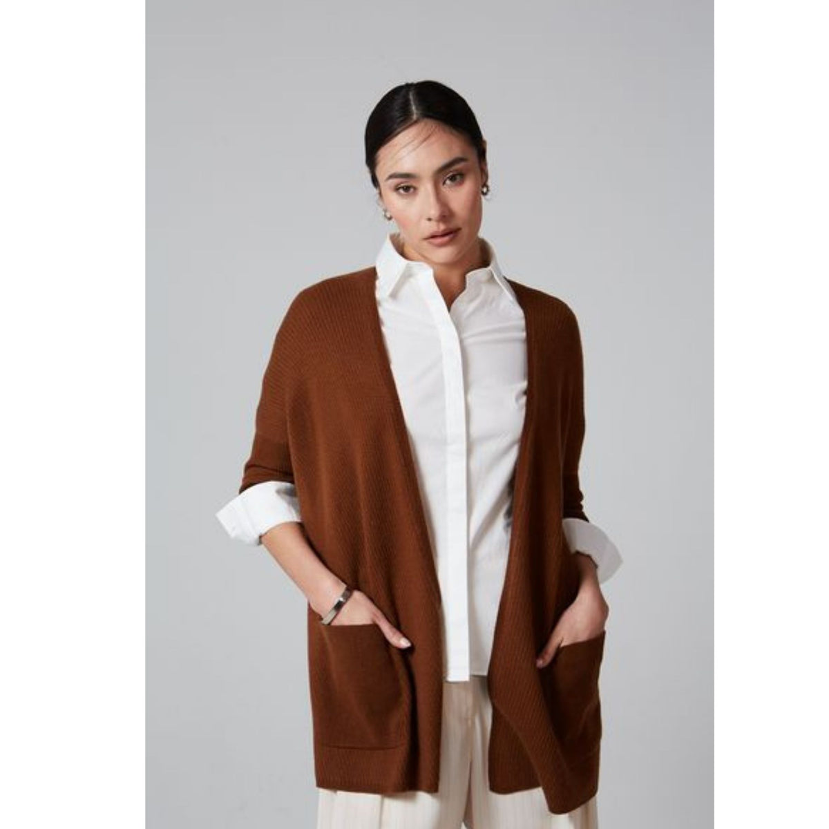 A woman stands against a gray background wearing the OO-TO Syrup Brown Lillien Open Cardigan, a handcrafted piece from Nepal, over a white button-up shirt and beige pants, with her hands in her cardigan pockets.