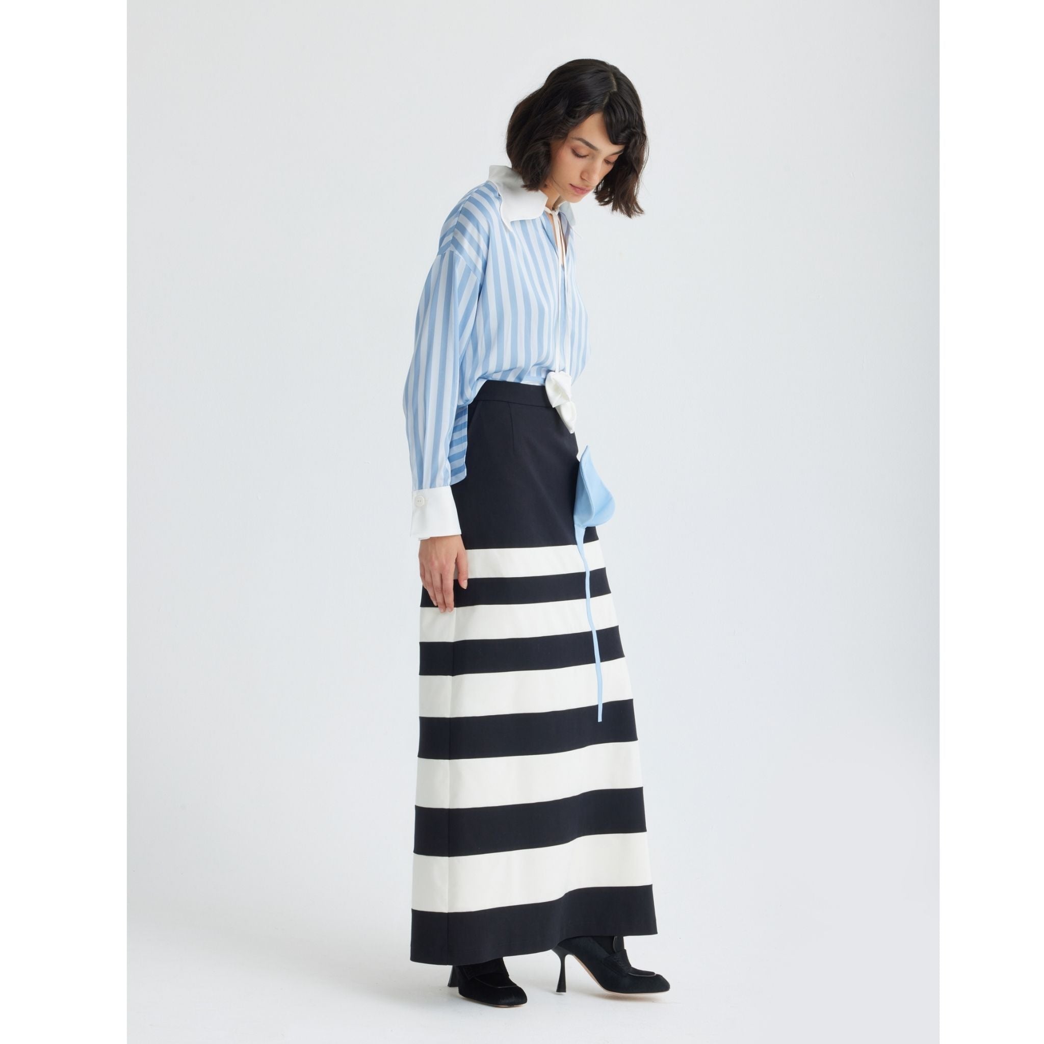 A woman with short dark hair wears JULIA ALLERT's Smart Elegance – Silk-Touch Striped Long-Sleeve Blouse in light blue, styled with a long black and white striped skirt and black heeled boots, standing against a white background.