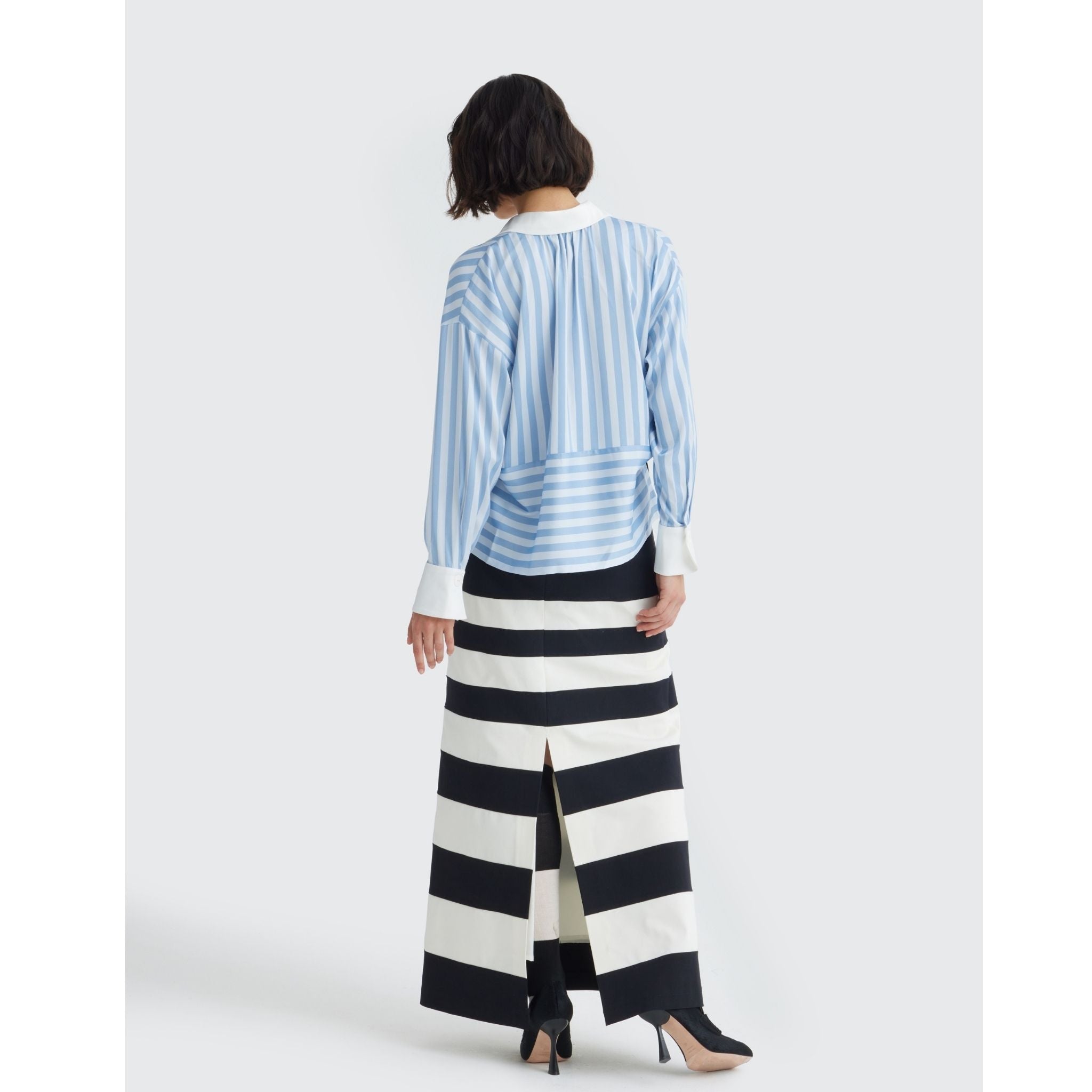 A woman faces away, wearing the JULIA ALLERT Smart Elegance Silk-Touch Striped Long-Sleeve Blouse in light blue with an oversized white collar, paired with a long black and white striped skirt and black heels.