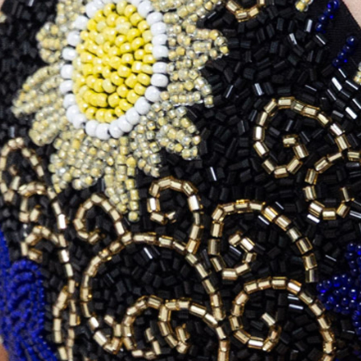 Close-up of the SELF-ADORE Dark Tide Hand-Beaded Embellished Bikini Top in black, featuring a yellow and white flower with a yellow center and swirling gold, black, and blue bead patterns on a black fabric background.