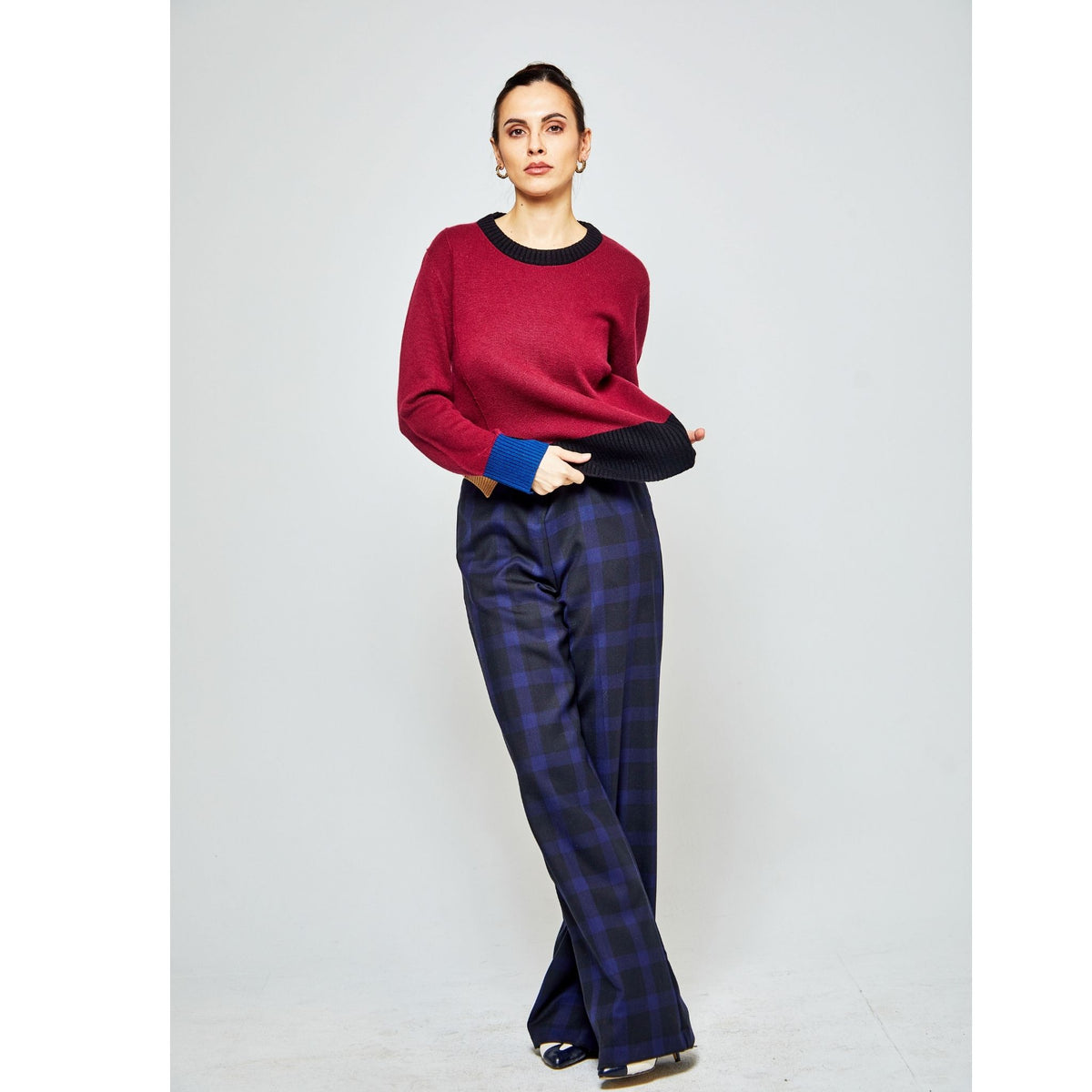 A woman stands against a plain light background, wearing the OO-TO Multi-tone Wine Onyx Crew Neck Sweater with black trim and blue cuffs, paired with loose-fitting navy and black checkered pants. She looks forward with a relaxed posture.