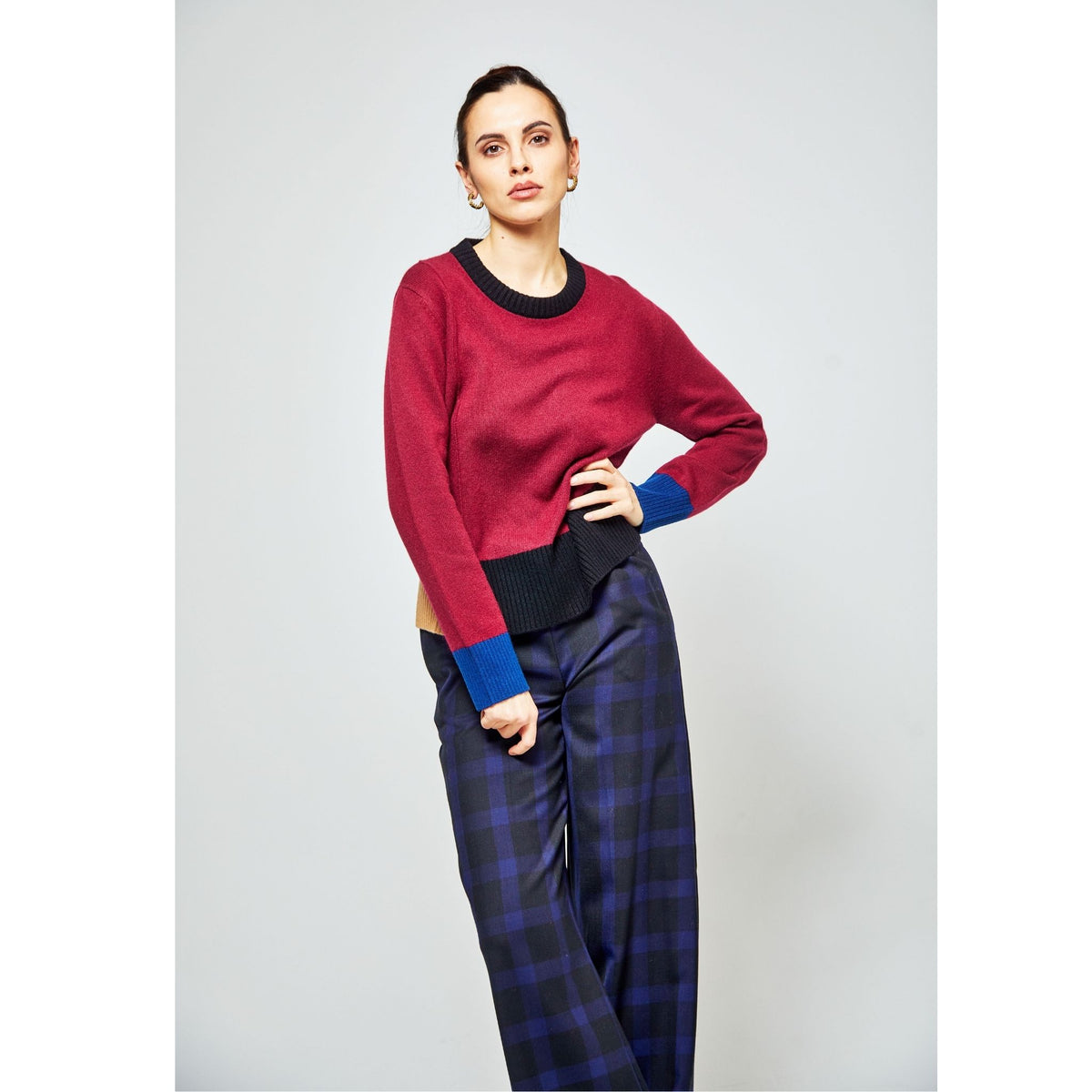 A woman stands confidently in an OO-TO Multi-tone Wine Onyx Crew Neck Sweater with blue cuffs and dark plaid wide-leg pants, posing with one hand on her hip against a plain light backdrop.