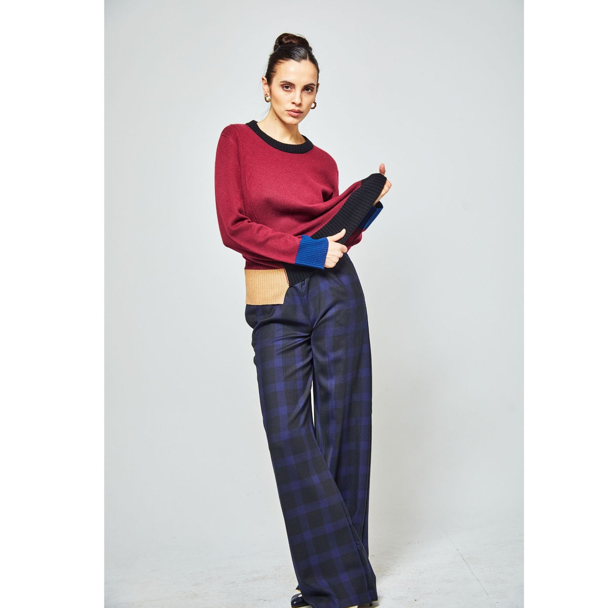 A woman poses in a studio wearing the OO-TO Multi-tone Wine Onyx Crew Neck Sweater with blue and tan cuffs, paired with high-waisted, wide-leg navy and black plaid pants against a light background.