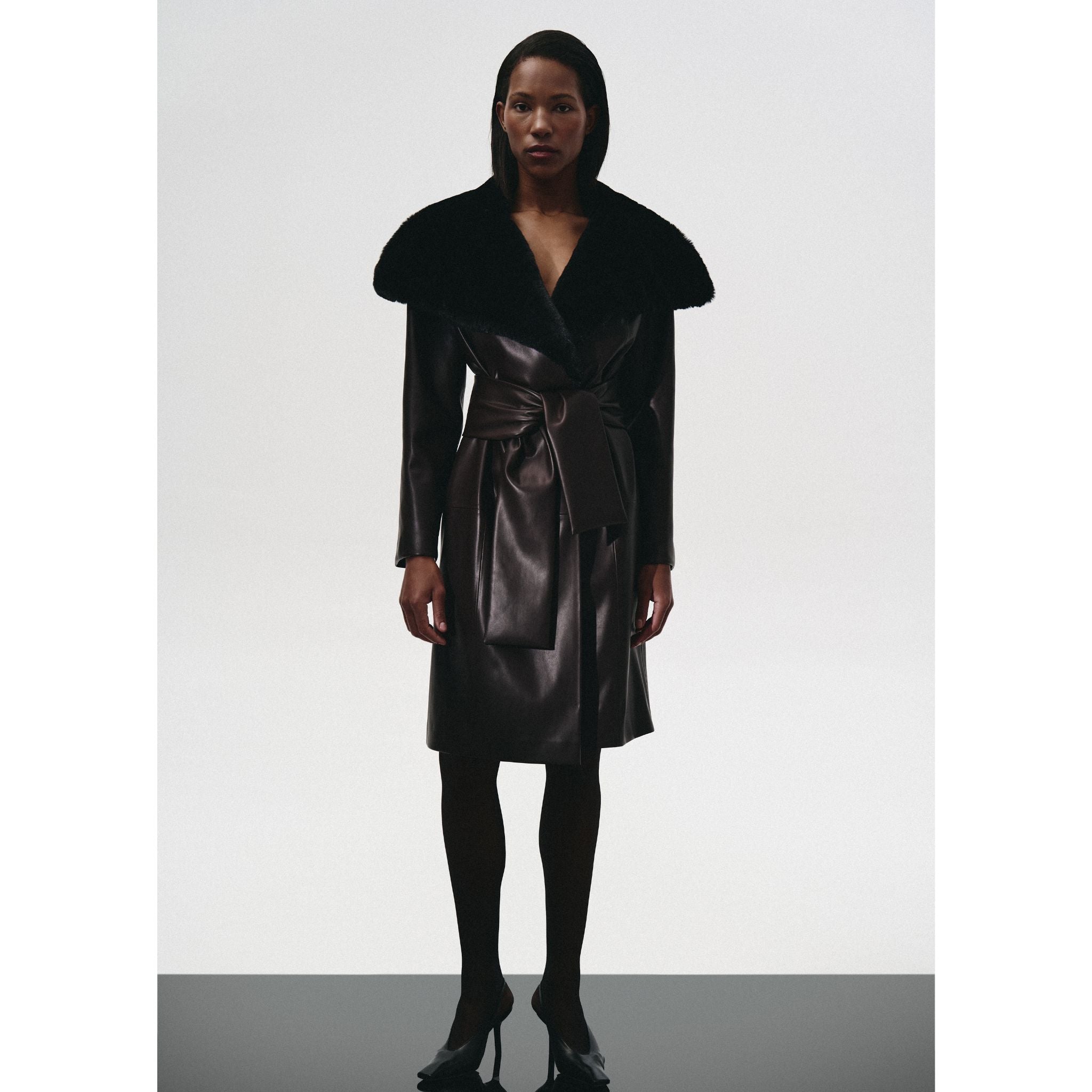 A woman faces forward in a KRIS MARÁN Leather Trench Coat With Fur Collar, detailed with a wide textured black fur collar and matching belt. She pairs it with dark tights and pointed black heels against a plain light background.