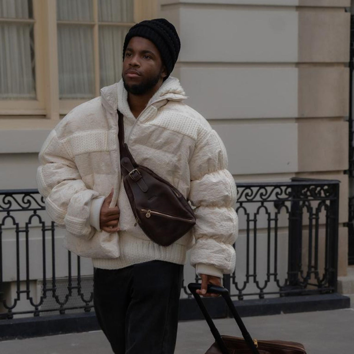 Person in a white textured jacket and black pants walking with a suitcase on a city street.