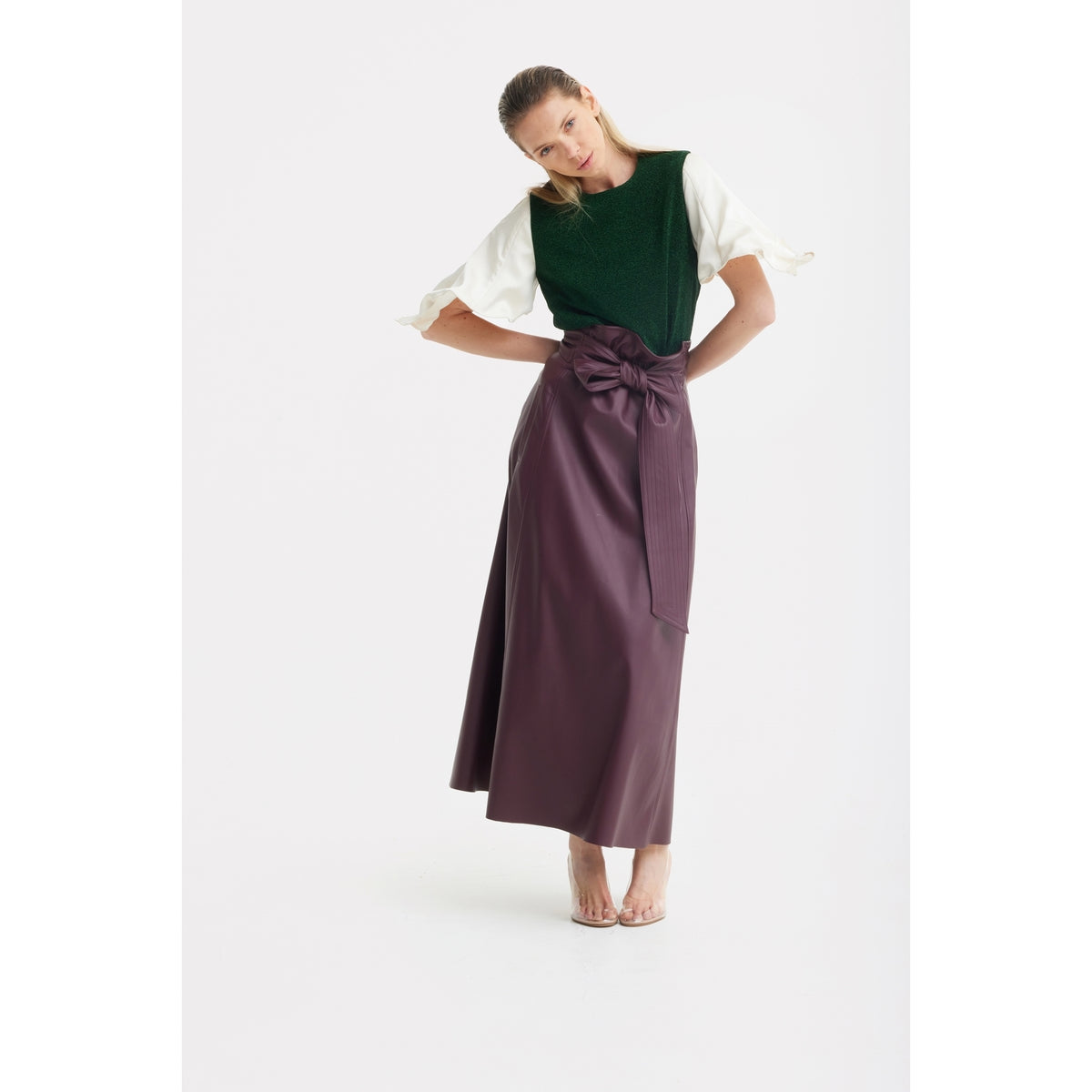 A woman models the JULIA ALLERT Green Lurex Knit Top With Adjustable Satin Sleeves, paired with a long mauve skirt featuring a bow, and clear open-toe heels, against a plain white background.