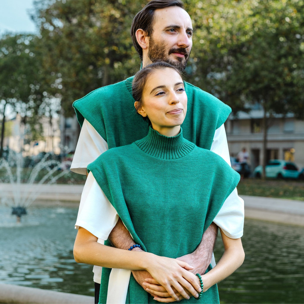 Two people wearing green and white outfits standing in a park with trees and a fountain in the background.