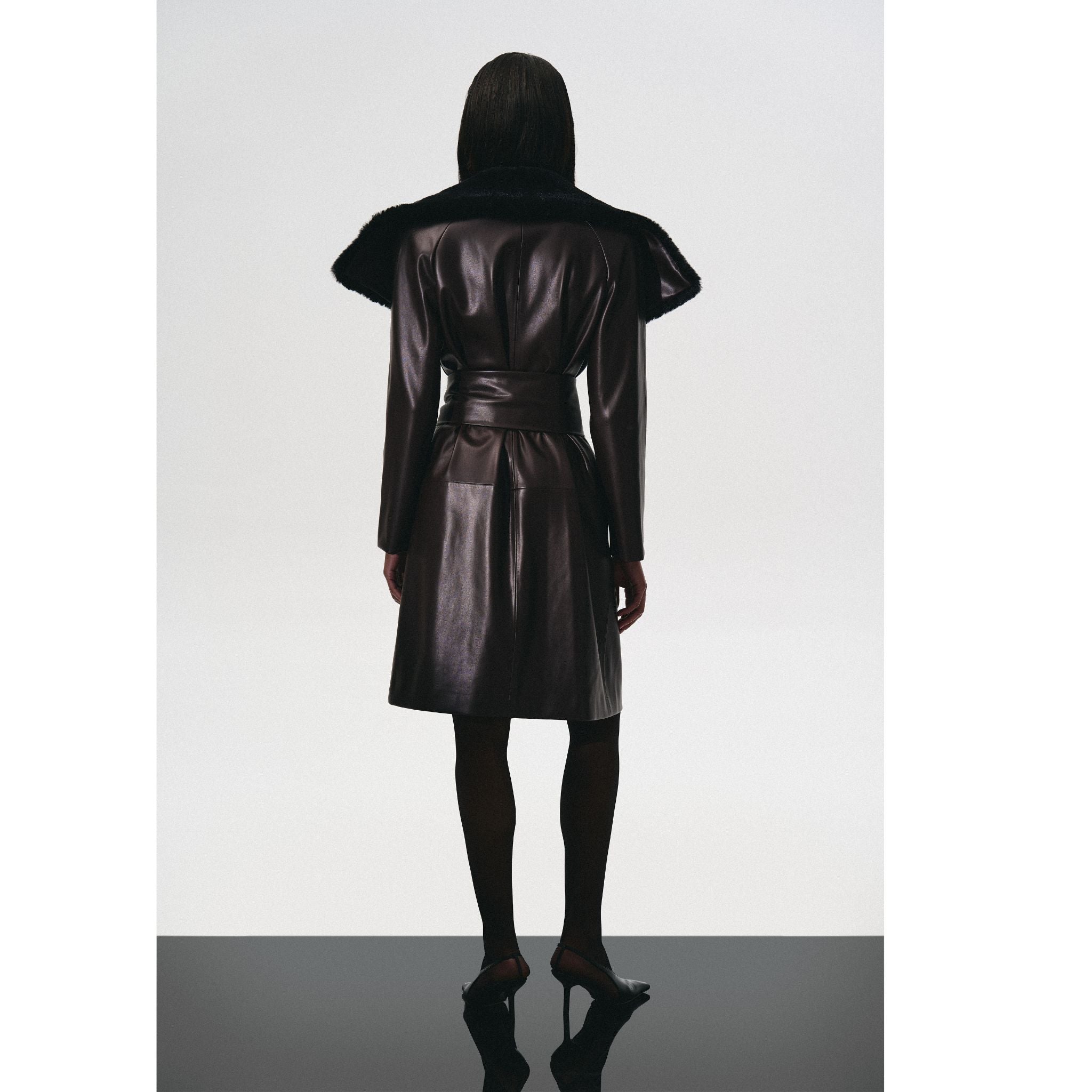 A person faces away, wearing the KRIS MARÁN Leather Trench Coat With Fur Collar, styled with black tights and heels, set against a plain light background.