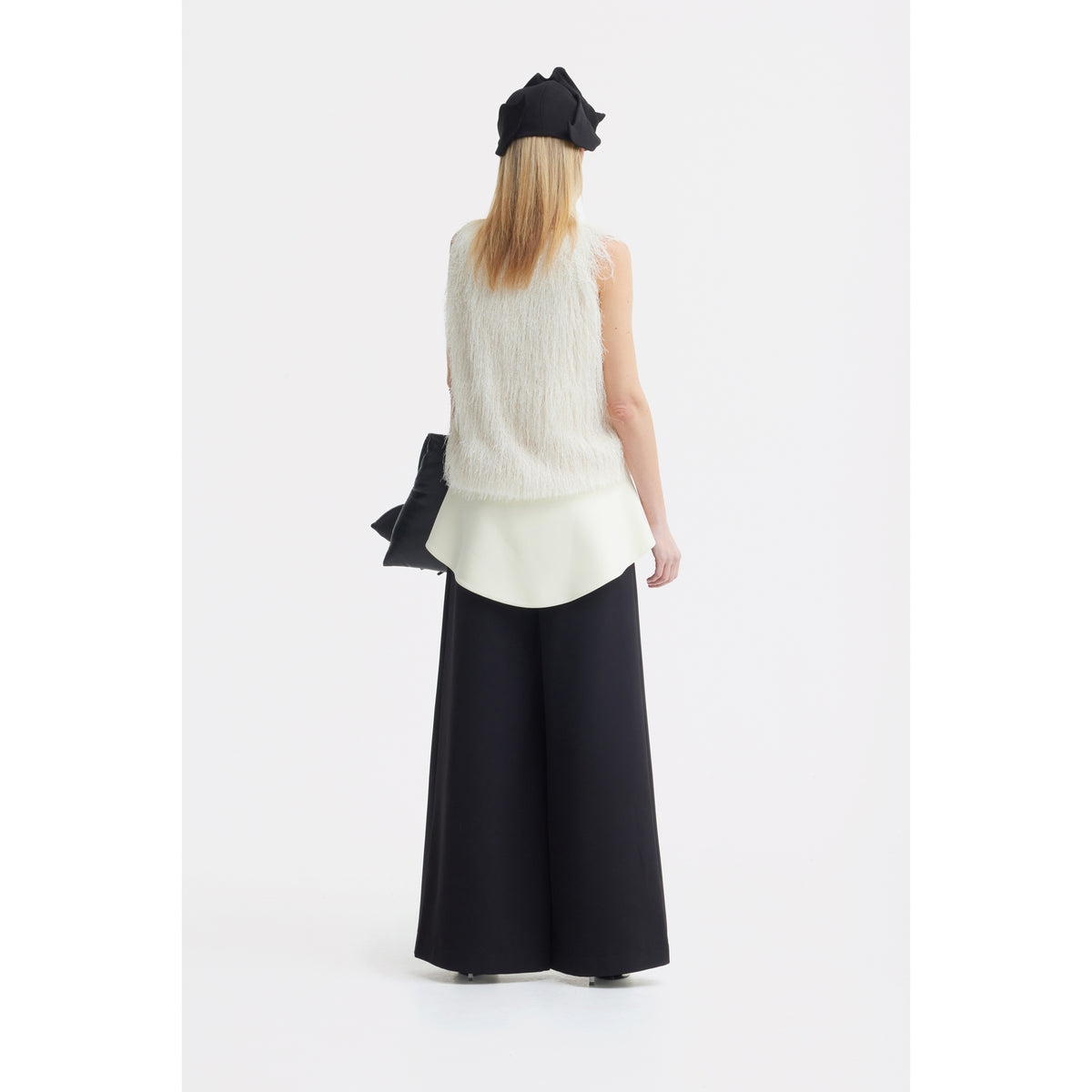 A person stands facing away against a plain white background, wearing a white sleeveless fuzzy top over a long white shirt, JULIA ALLERT Statement Waist Wide-Leg Trousers in black, and a black hat.