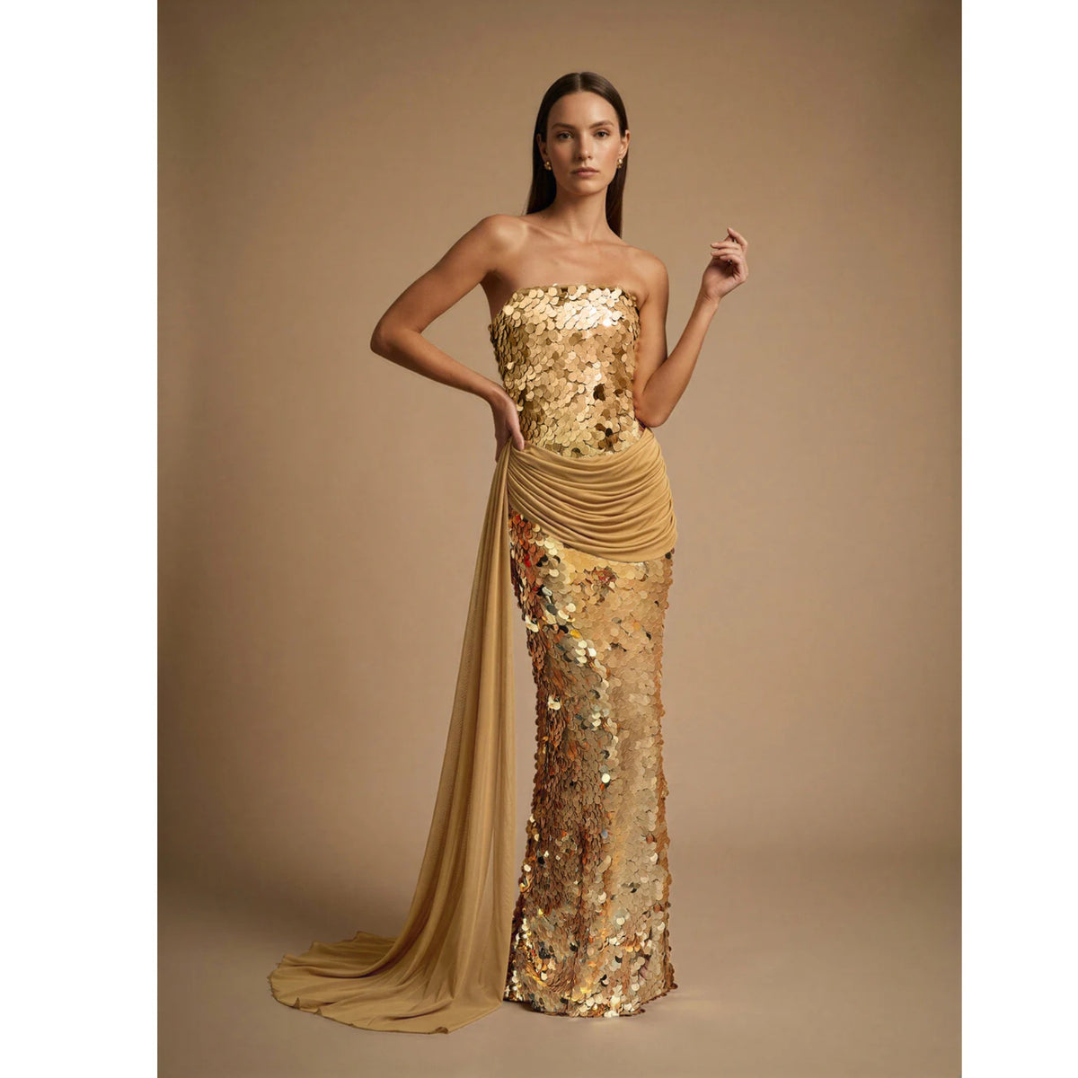 A woman poses confidently against a beige background in the ASHLEY STAMBOULI Alana Dress | PR Sample—a strapless, mermaid gown with gold sequins and a draped sheer sash, one hand on her hip and the other raised.