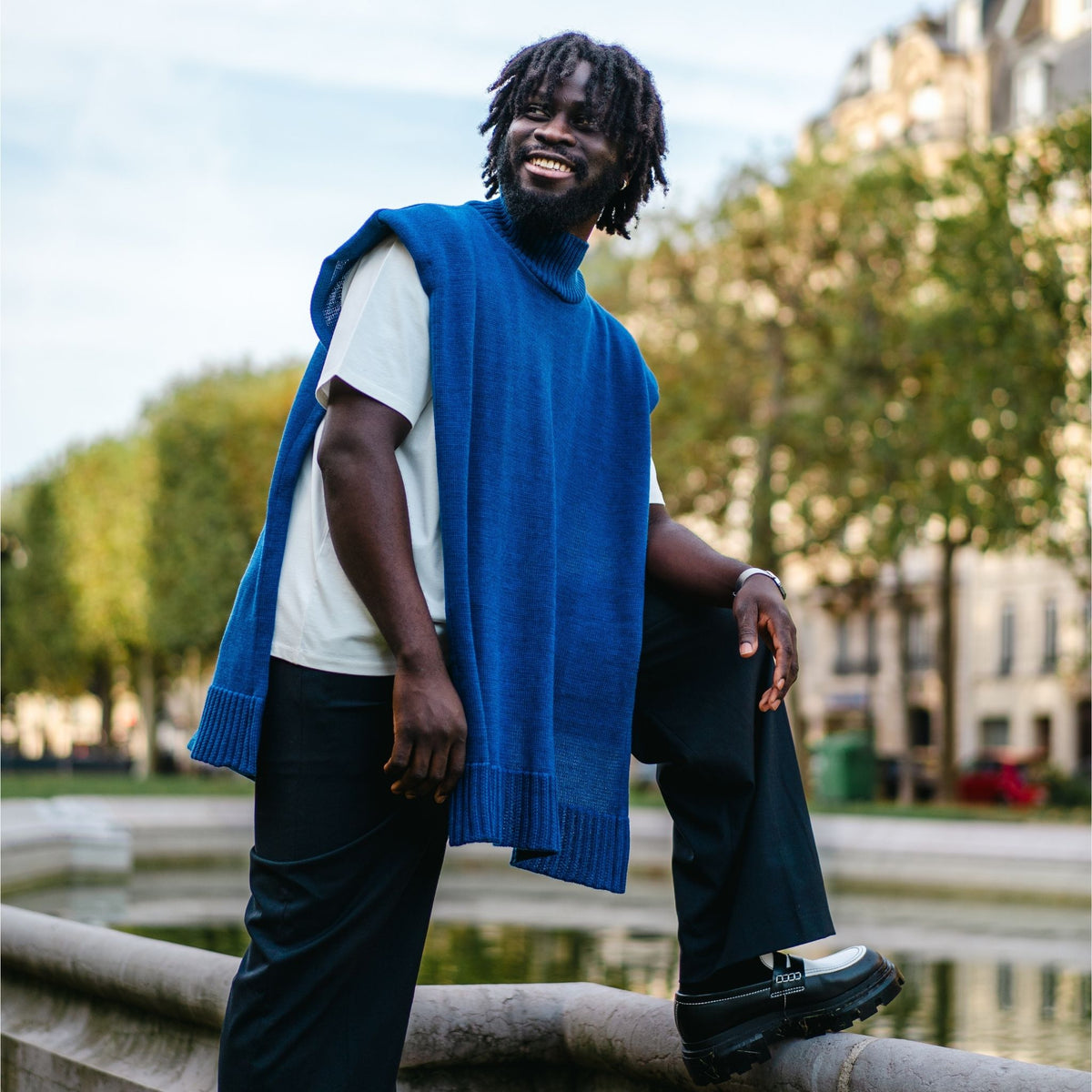 Man wearing a blue sweater vest sitting on a stone ledge with trees and buildings in the background