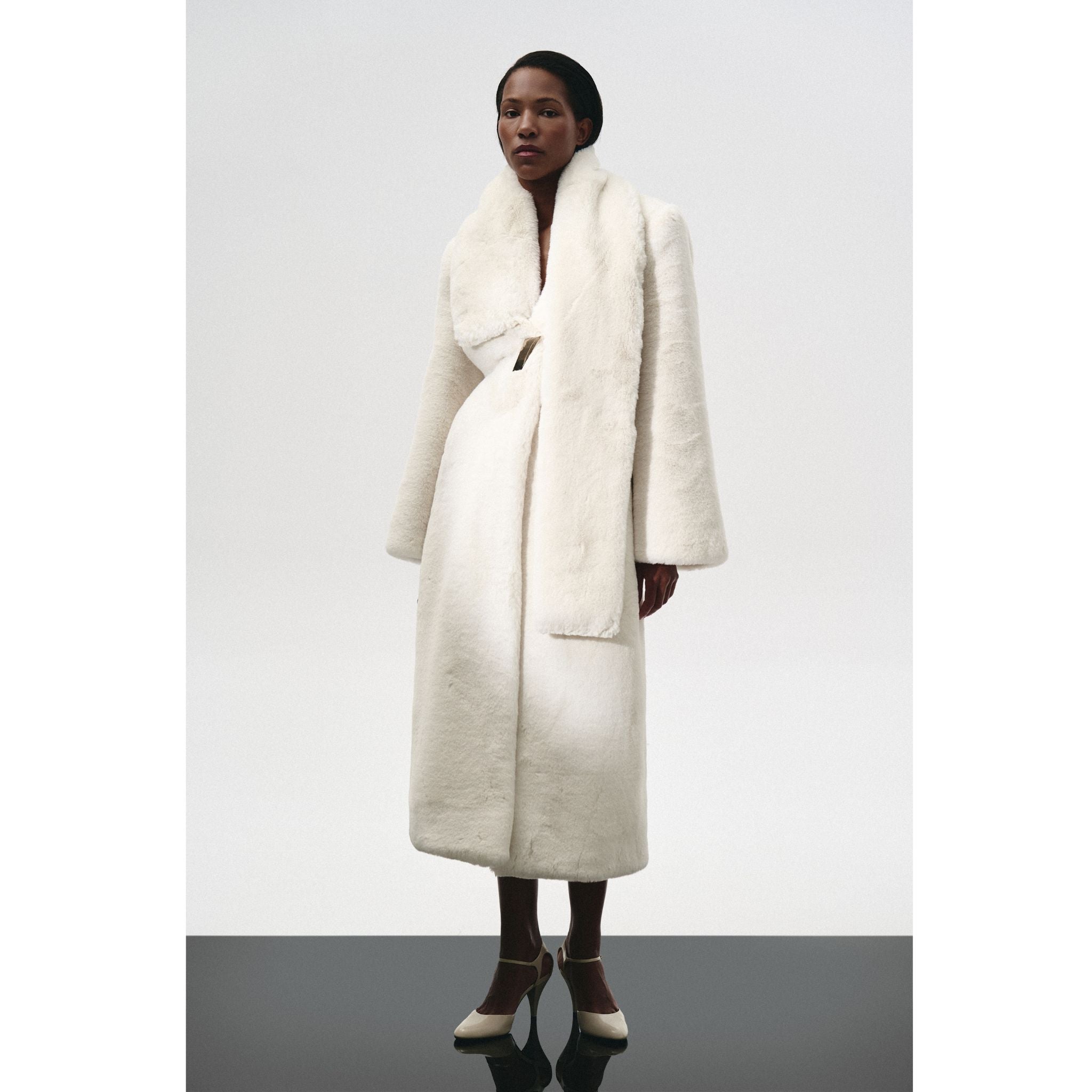 A person stands against a plain background wearing the KRIS MARÁN Fur Maxi Coat With Shawl Collar, styled with white ankle-strap heels.