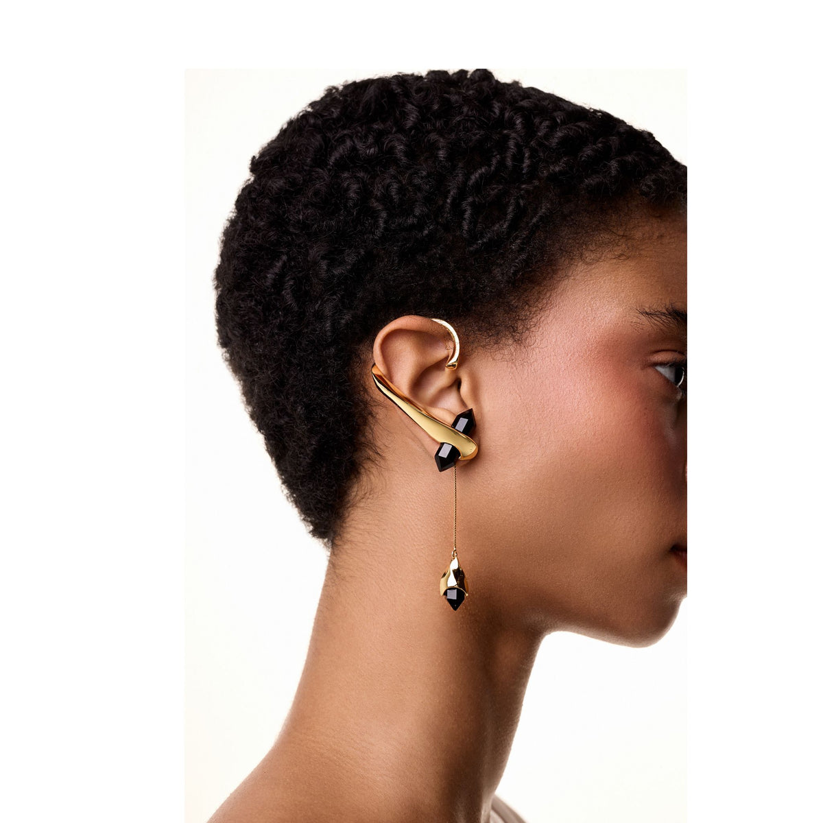 A woman with short, curly hair wears the ANNA PRATA Gold Dualis Earcuff from the Corpo E Alma Collection, featuring sculptural gold and black onyx accents that extend from the upper ear and dangle a matching drop. The background is white.