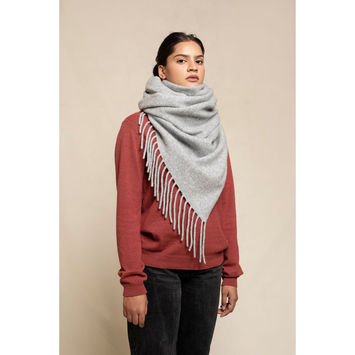 A woman in a red sweater and black pants stands facing forward, wearing the OO-TO Grey Knitted Felted Cashmere Chunky Scarf wrapped around her neck and shoulders. The background is plain beige.
