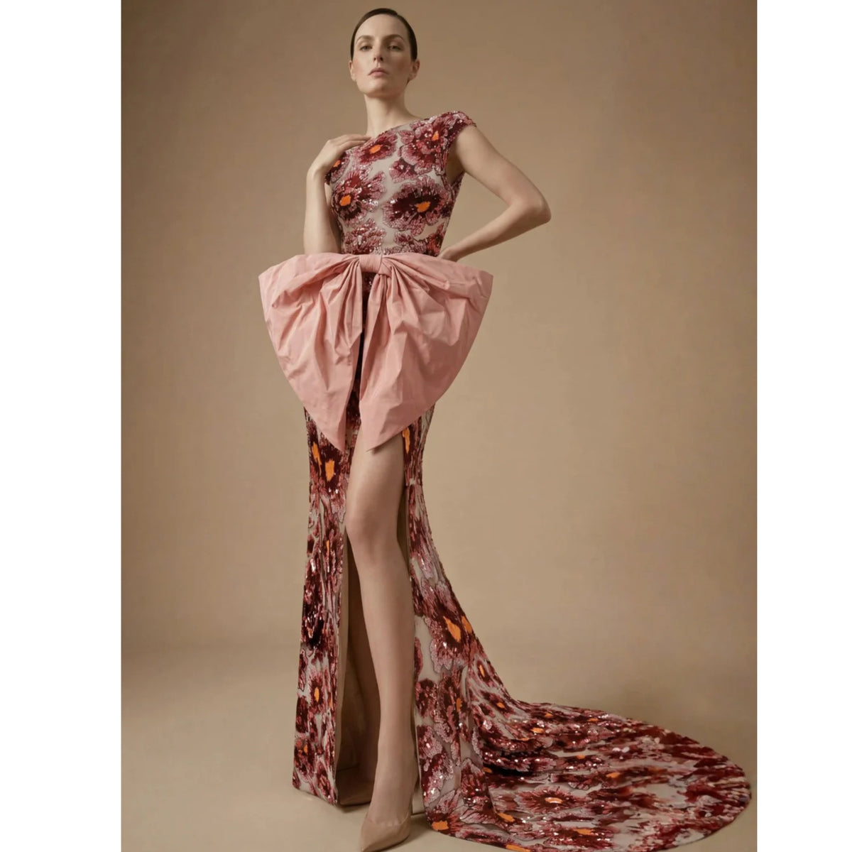 A woman poses confidently against a beige backdrop in the Coralia Dress by ASHLEY STAMBOULI, a floral red and pink evening gown with a dramatic high slit, large pink bow at the waist, and a long train. PR Sample.