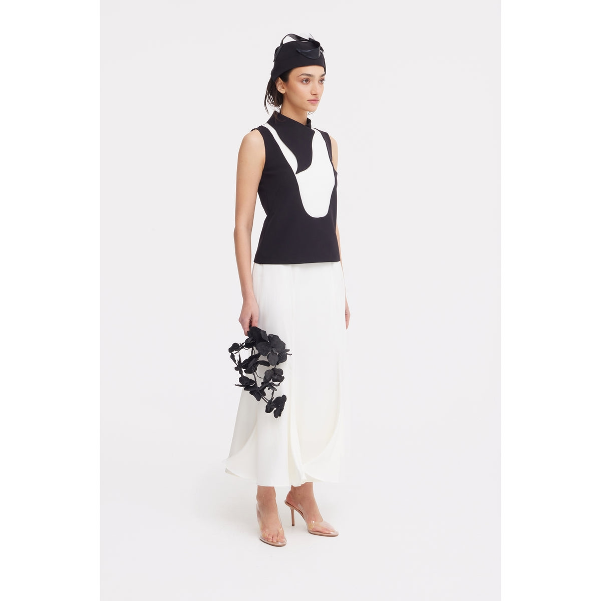 A woman poses against a white background in JULIA ALLERT eveningwear, pairing a black sleeveless top with a white panel and the High Waist Double-Sided Satin Maxi Skirt in Ivory, nude heels, a black headpiece, and holding a black floral accessory.