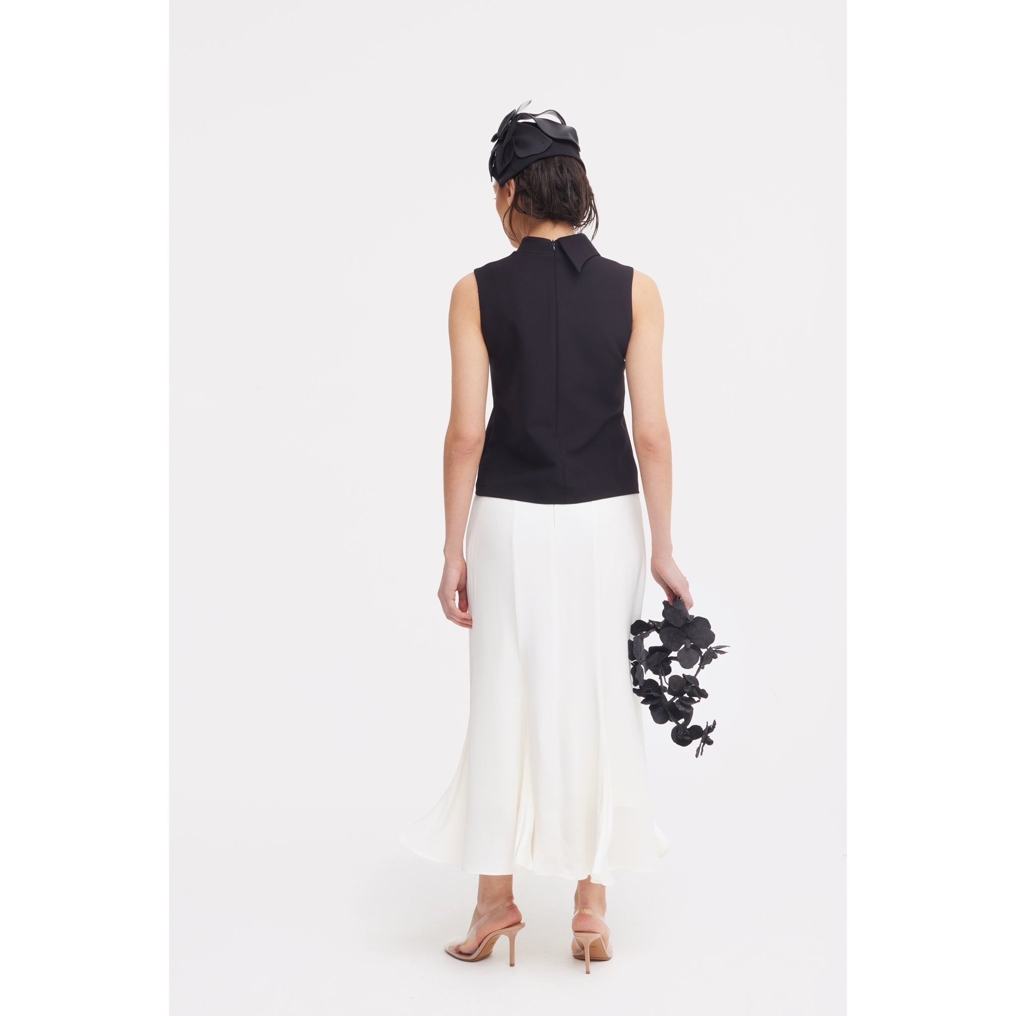 A woman faces away, wearing a sleeveless black top, JULIA ALLERT High Waist Double-Sided Satin Maxi Skirt in ivory, nude heels, and a black fascinator. She holds black floral décor against a plain white background—ideal for eveningwear.
