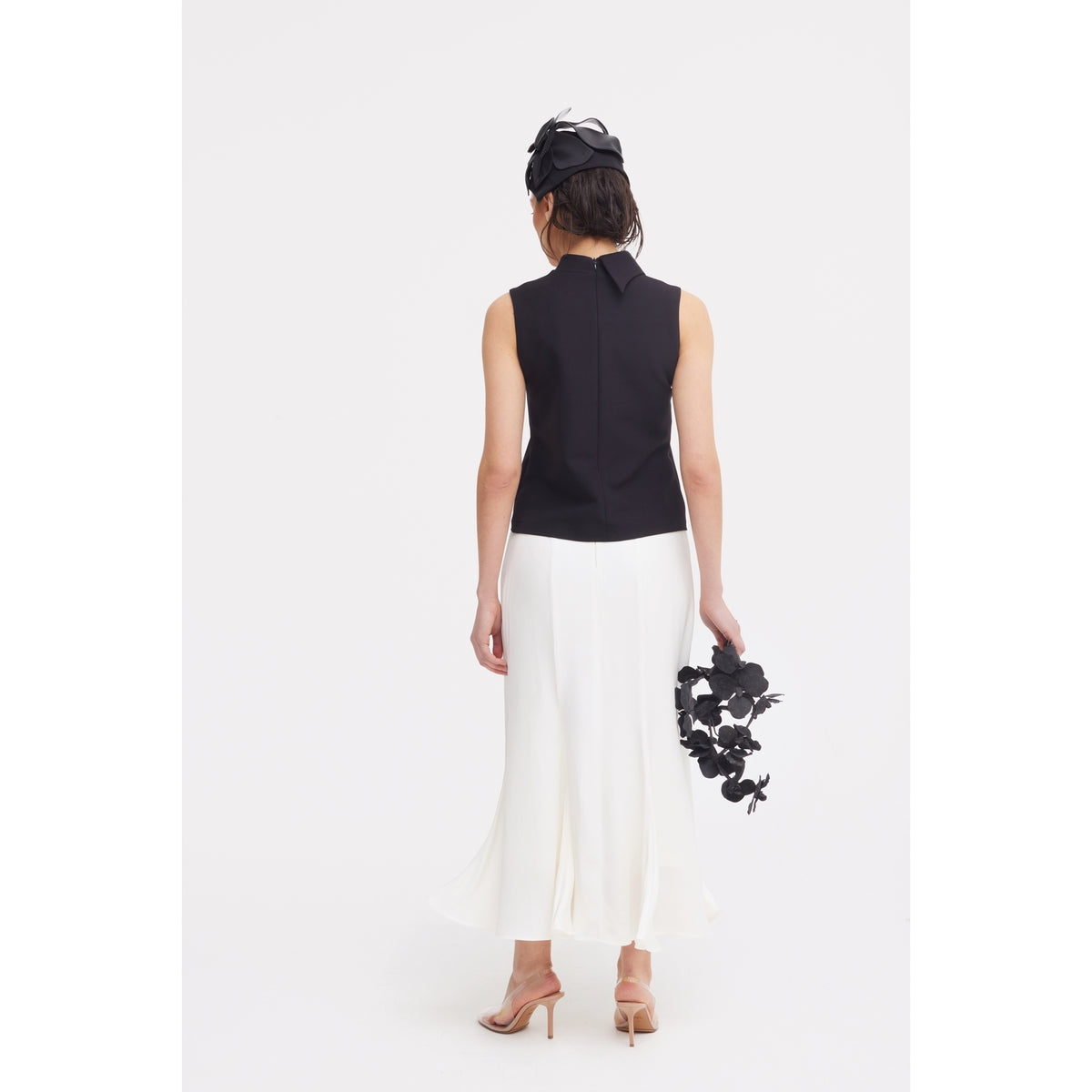 A woman faces away, wearing a sleeveless black top, JULIA ALLERT High Waist Double-Sided Satin Maxi Skirt in ivory, nude heels, and a black fascinator. She holds black floral décor against a plain white background—ideal for eveningwear.