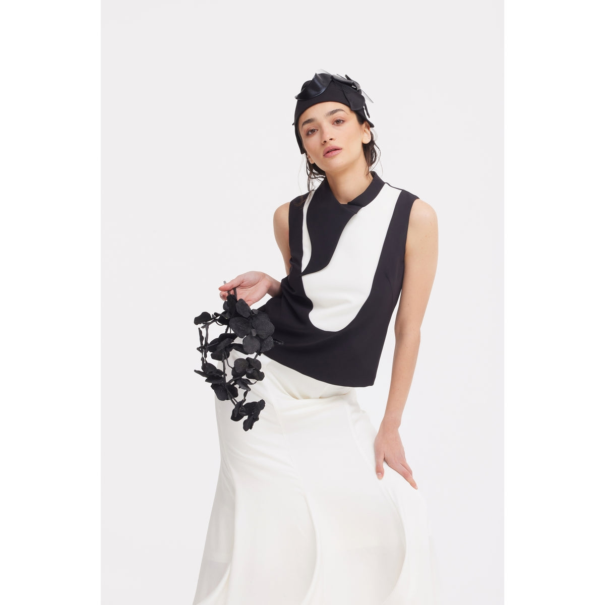 A woman in a JULIA ALLERT black and white sleeveless top, High Waist Double-Sided Satin Maxi Skirt - Ivory, and a black headpiece poses gracefully, holding black artificial flowers against a plain white background.
