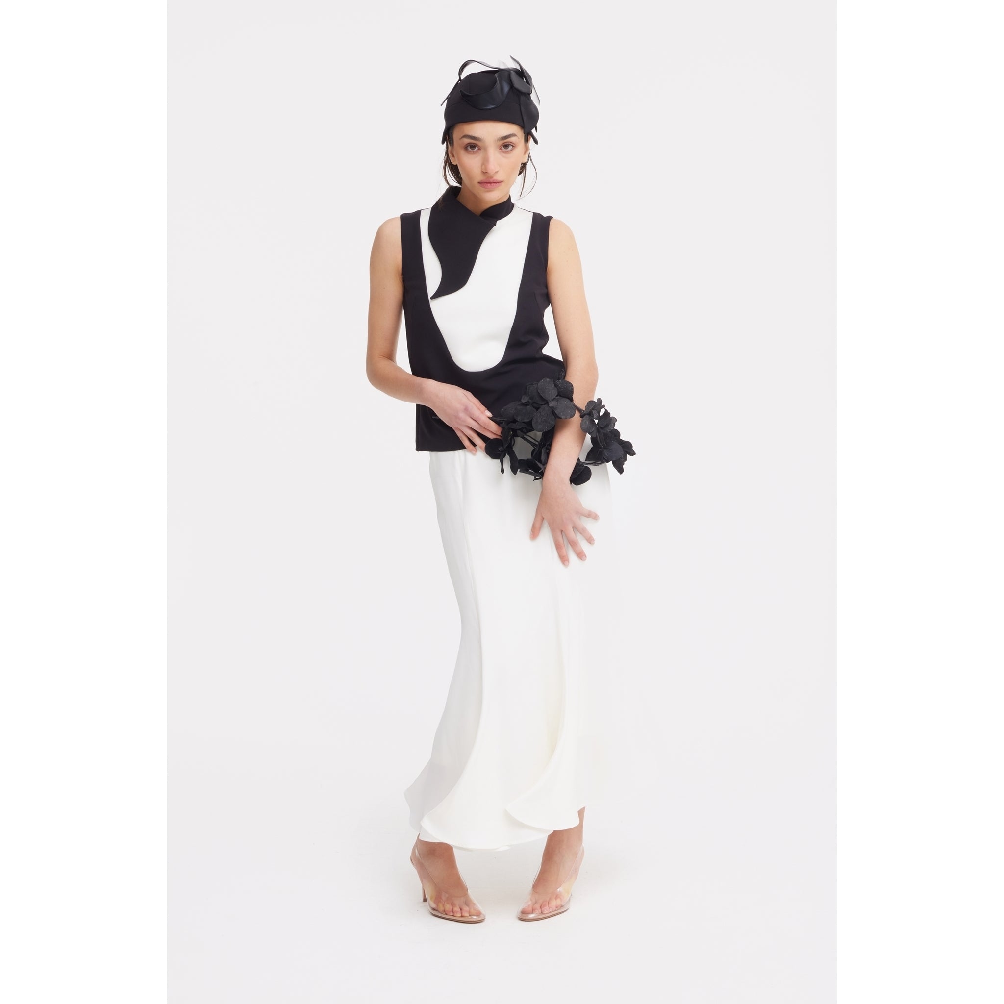 A woman poses in a sleeveless black and white top, JULIA ALLERT High Waist Double-Sided Satin Maxi Skirt - Ivory, clear heels, and a black headpiece, holding a black floral accessory against a plain white background—ideal for chic evenings.