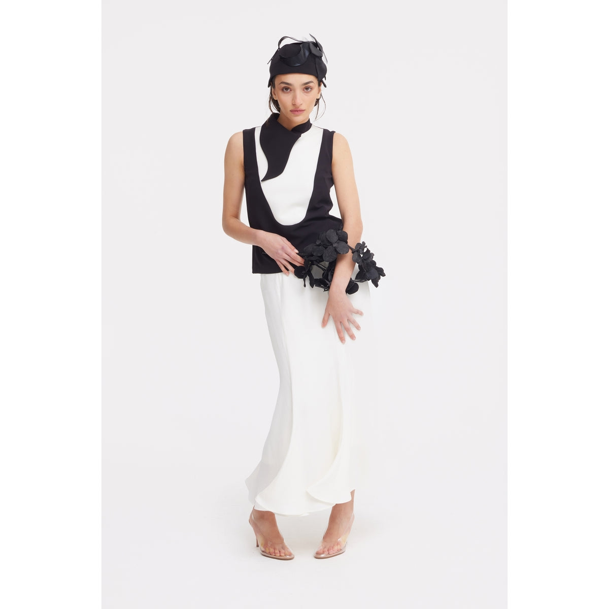 A woman poses in a sleeveless black and white top, JULIA ALLERT High Waist Double-Sided Satin Maxi Skirt - Ivory, clear heels, and a black headpiece, holding a black floral accessory against a plain white background—ideal for chic evenings.