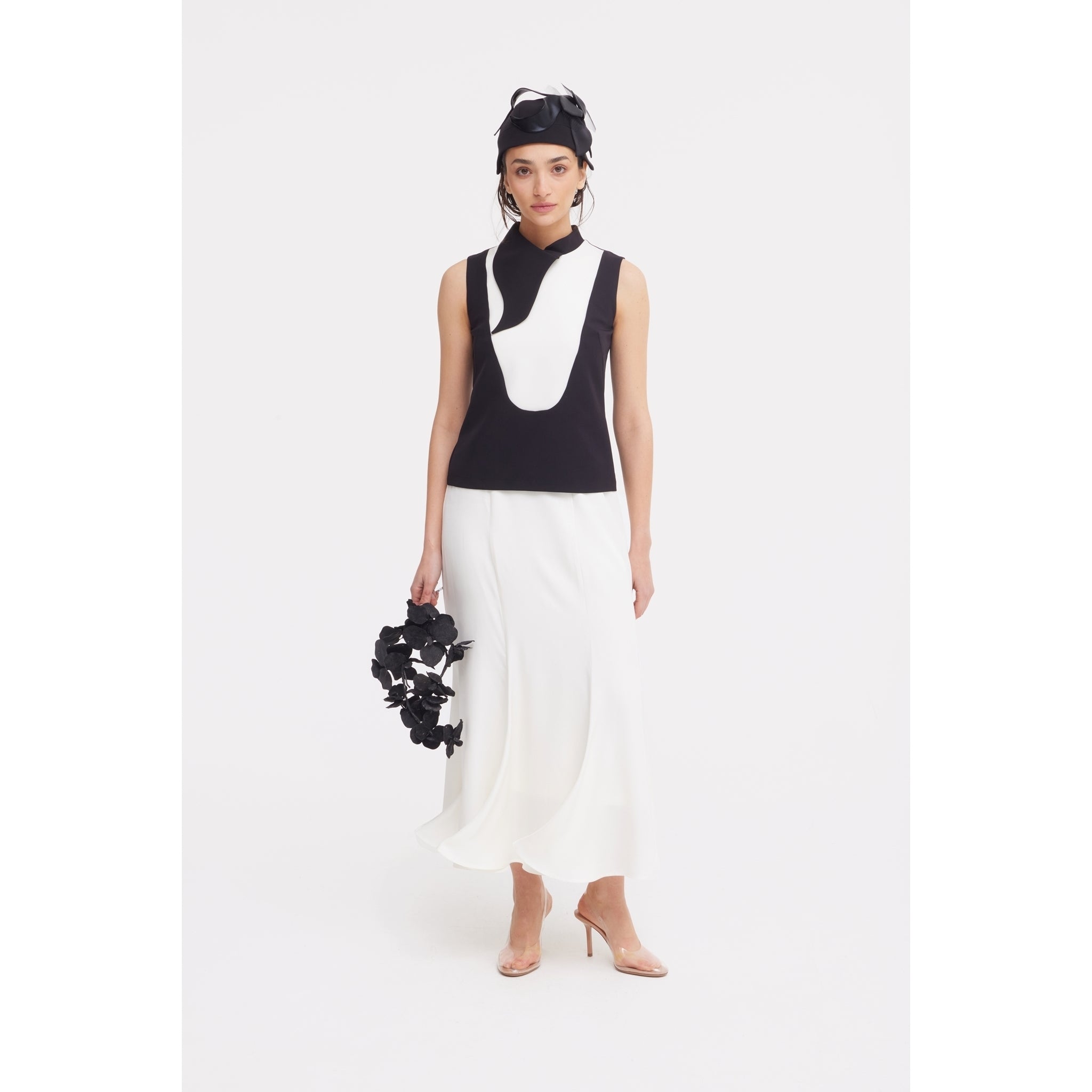 A woman wears a sleeveless black and white top, JULIA ALLERT High Waist Double-Sided Satin Maxi Skirt in Ivory, nude heels, and a black headpiece. Holding a black floral accessory and standing against a white background—elegant eveningwear.