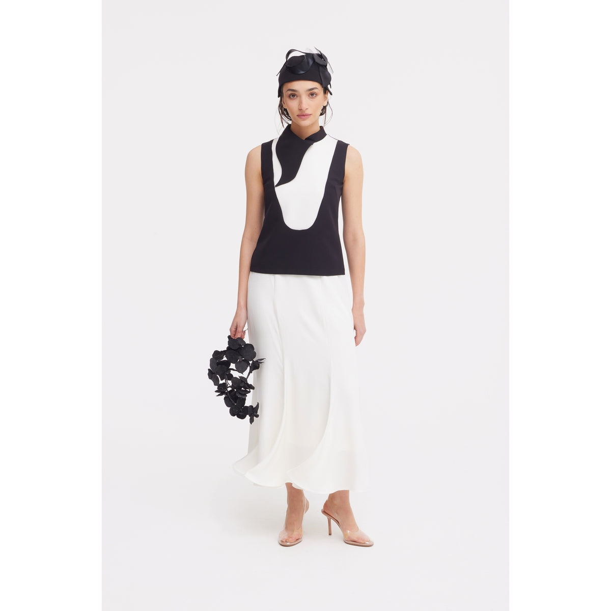 A woman wears a sleeveless black and white top, JULIA ALLERT High Waist Double-Sided Satin Maxi Skirt in Ivory, nude heels, and a black headpiece. Holding a black floral accessory and standing against a white background—elegant eveningwear.