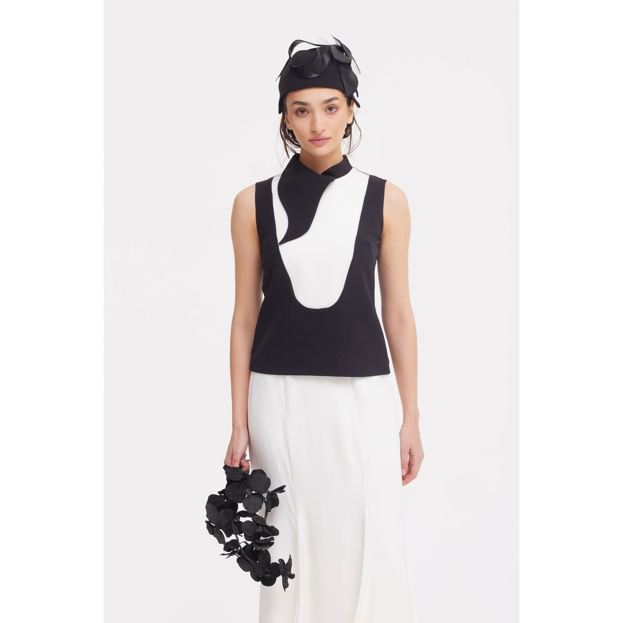 A woman poses against a white background wearing a sleeveless black and white top with abstract designs, the JULIA ALLERT High Waist Double-Sided Satin Maxi Skirt in Ivory, a black headpiece, and holding a black floral accessory for an elegant look.