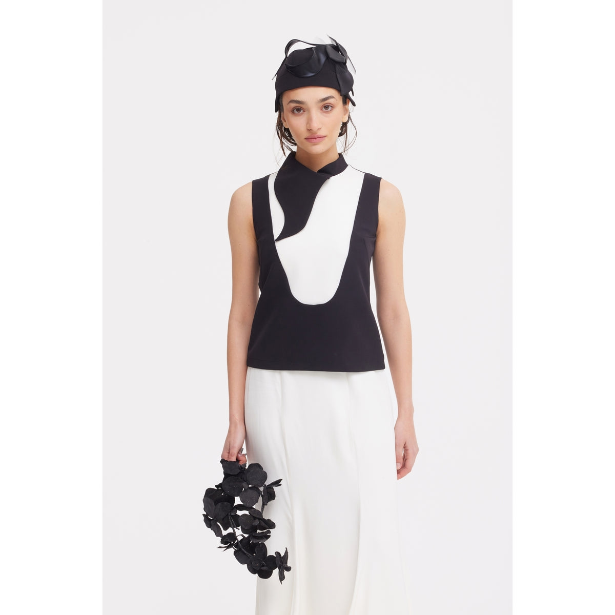 A woman poses against a white background wearing a sleeveless black and white top with abstract designs, the JULIA ALLERT High Waist Double-Sided Satin Maxi Skirt in Ivory, a black headpiece, and holding a black floral accessory for an elegant look.