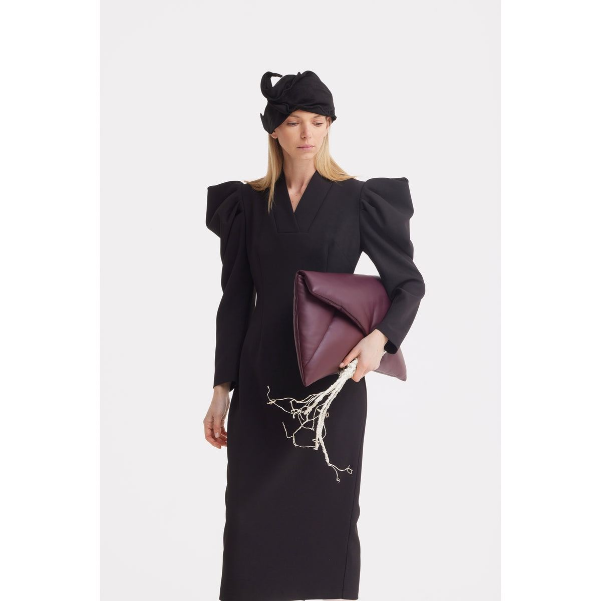 A woman in a black dress and the JULIA ALLERT Headpiece – Evening Mini Hat Style (Black) holds a large folded burgundy clutch and white branches against a plain white background.