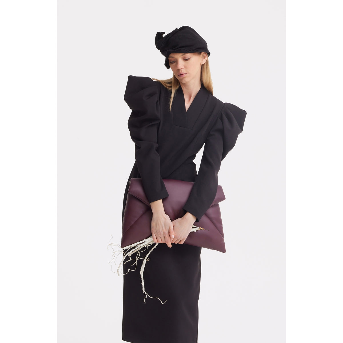 A woman wears a black JULIA ALLERT Headpiece - Evening Mini Hat Style and a black dress with bold, puffy shoulders, holding a burgundy envelope clutch among white twigs against a plain white background.