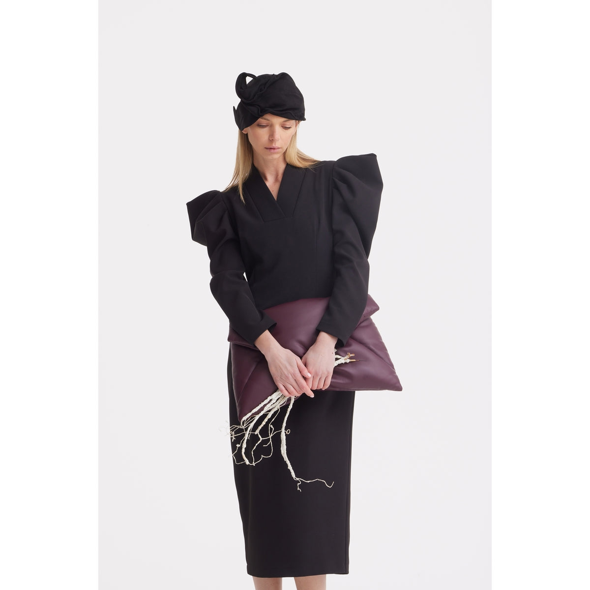 A woman in a black dress with puffed shoulders wears the JULIA ALLERT Headpiece - Evening Mini Hat Style - Black and holds a large burgundy clutch with exposed white branches against a plain white background.