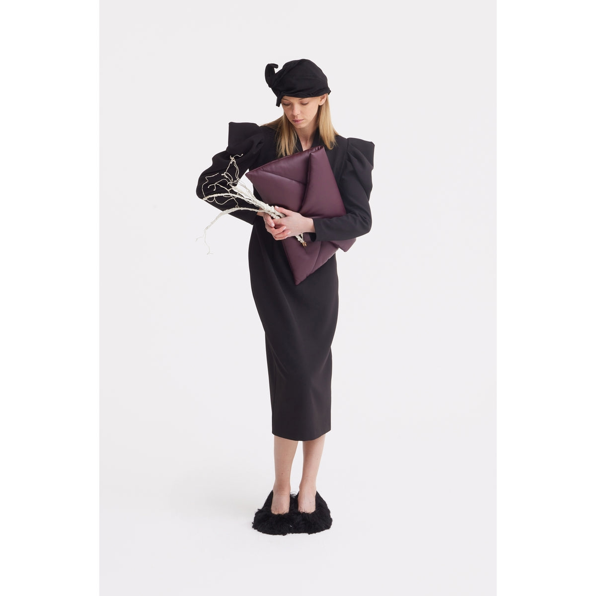 A woman in a black dress, sculptural JULIA ALLERT Evening Mini Hat Style headpiece in black, and black fuzzy shoes holds a large maroon clutch and white decorative branches against a plain white background.