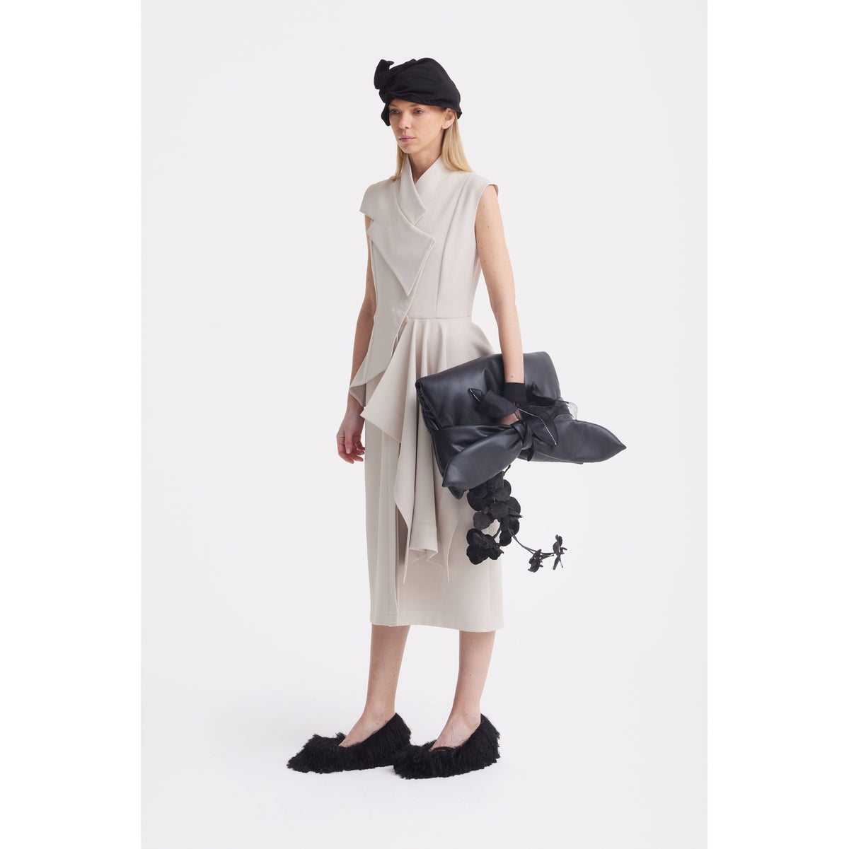 A woman stands against a white background wearing the JULIA ALLERT Architectural Draped Midi Dress in beige, styled with a black hat, black furry shoes, and a large black bag decorated with oversized bows and black flower details.