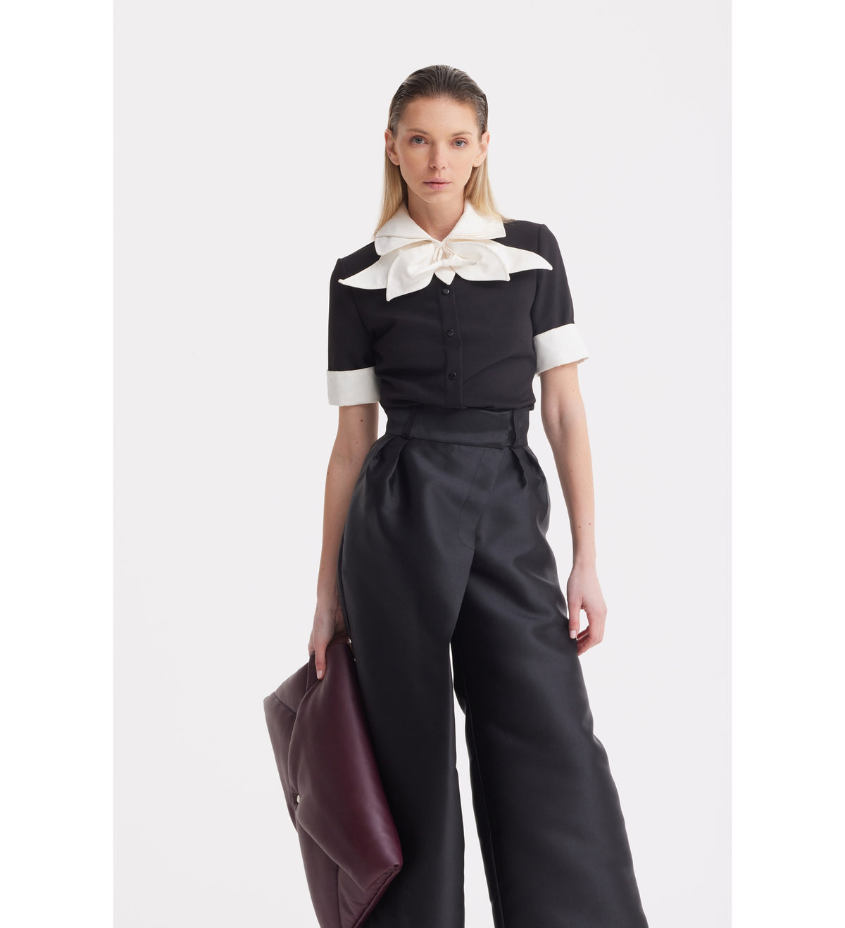 A woman stands against a white background wearing the JULIA ALLERT Short Sleeve Formal Bow Blouse in black, paired with high-waisted black wide-leg pants and holding a large dark burgundy clutch.