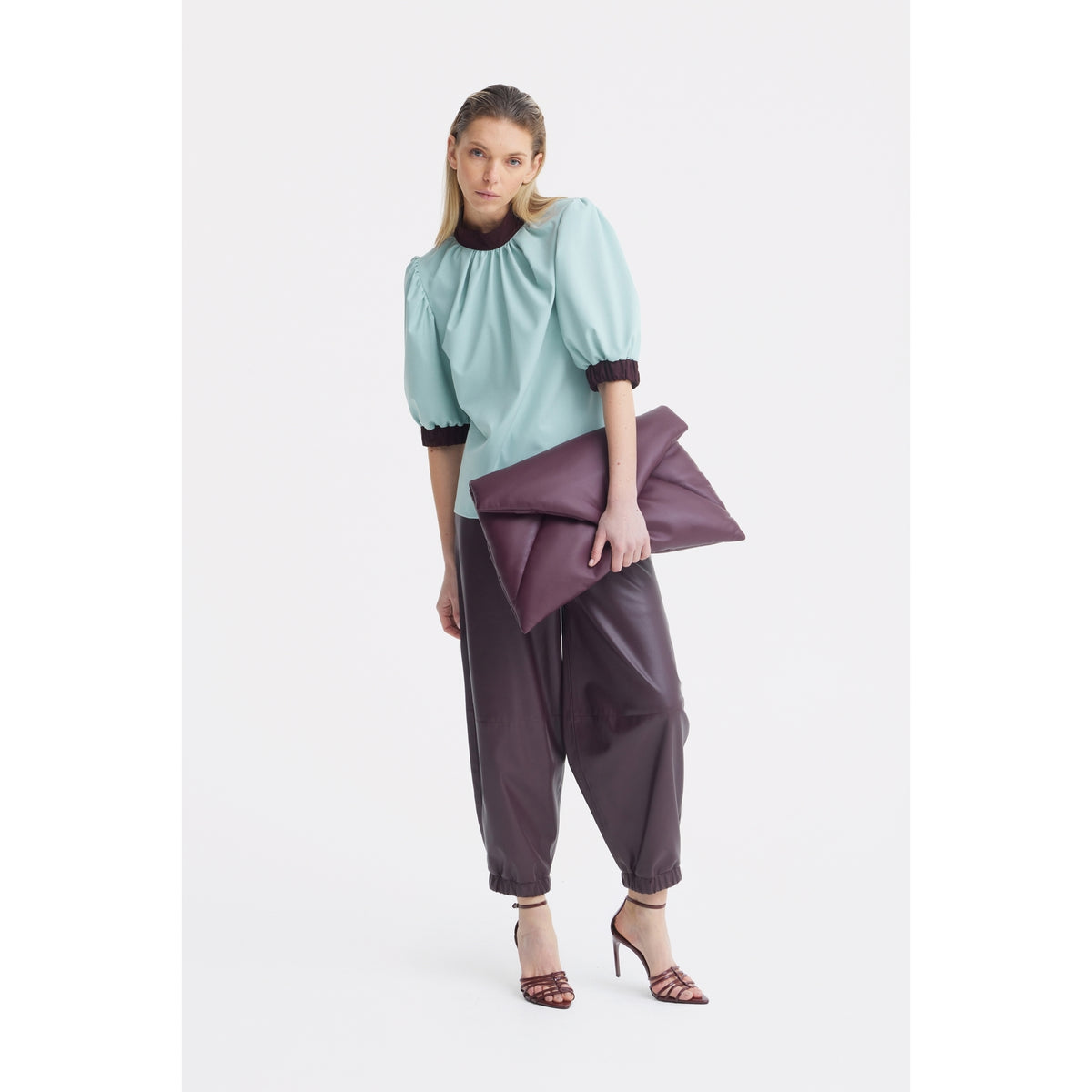 A woman stands against a white background wearing the JULIA ALLERT Textured Short Sleeve Blouse in light blue, paired with loose dark brown pants, strappy heels, and holding an oversized folded maroon clutch bag.