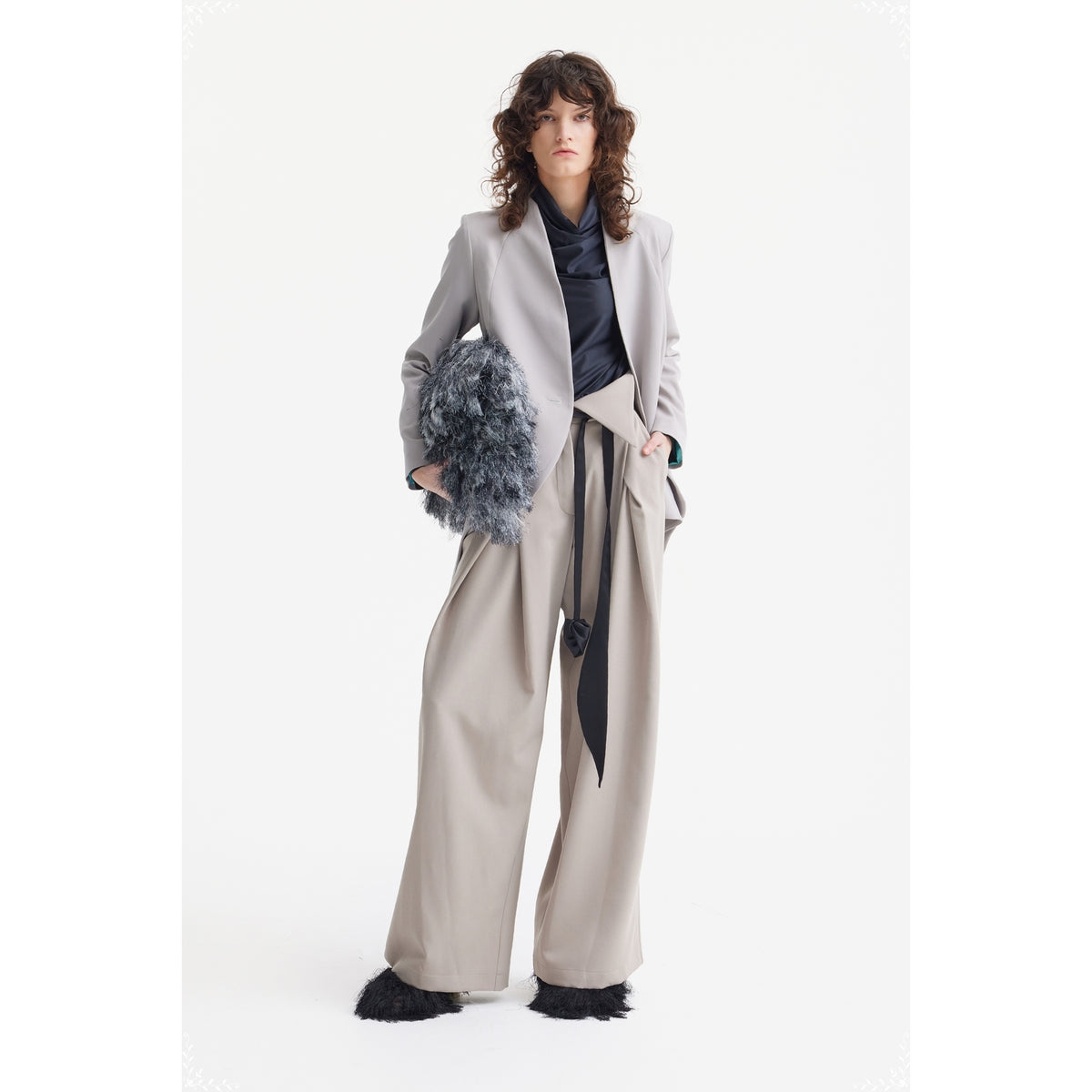 A person with curly hair wears a light gray blazer, a black top, JULIA ALLERT Fashion Forward Wide-Leg Pants &amp; Contrast Belt in beige, black furry shoes, and holds a large textured gray clutch against a white background.