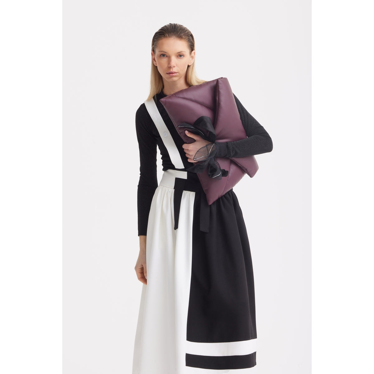 A woman with long hair wears the JULIA ALLERT Midi Apron Dress With Adjustable Straps - Black Ivory and a black top, holding a large dark purple clutch with a black ribbon against a plain white background.