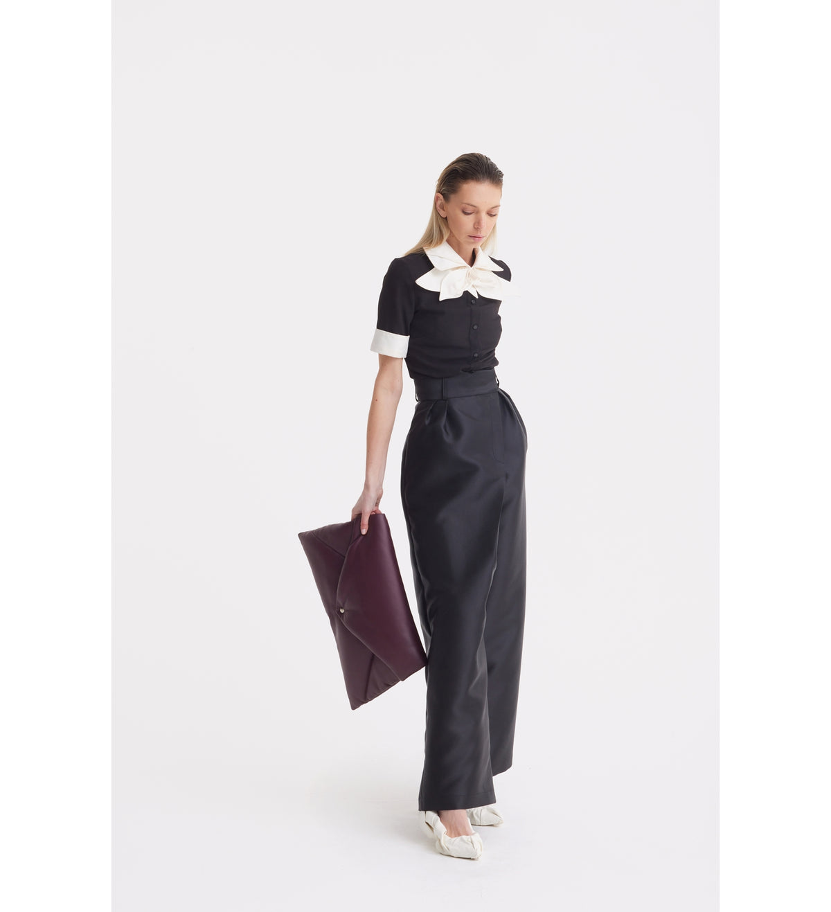A woman stands against a white background in the JULIA ALLERT Short Sleeve Formal Bow Blouse - Black, paired with high-waisted black trousers, white heels, and a large burgundy clutch. She looks downward with a neutral expression.
