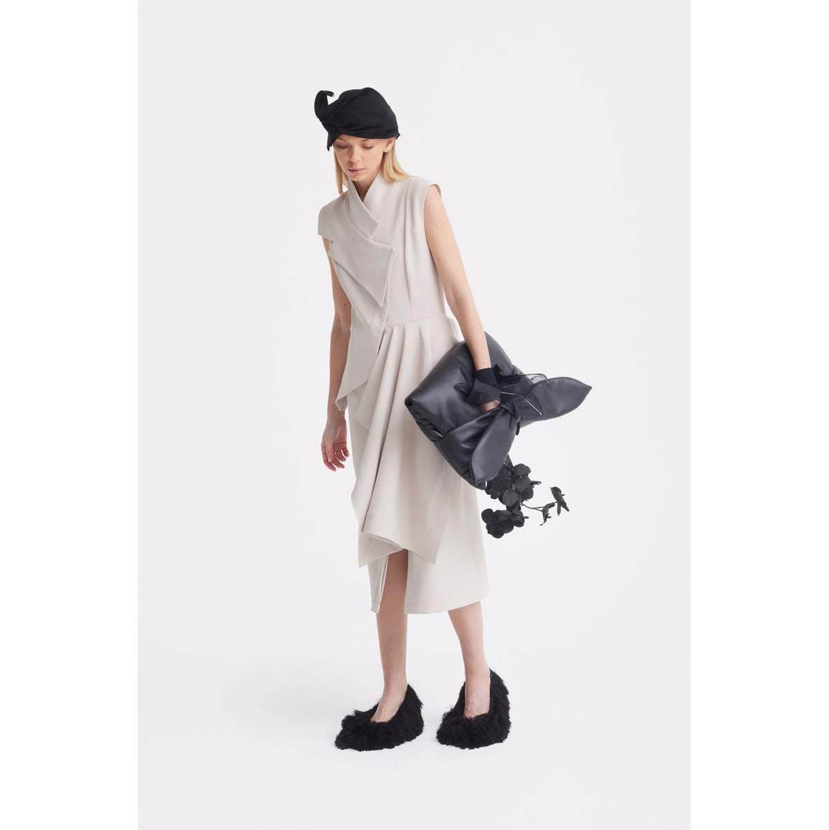 A woman in a JULIA ALLERT Architectural Draped Midi Dress - Beige, black furry shoes, and a black beret holds a large black handbag with flower details against a plain white background.