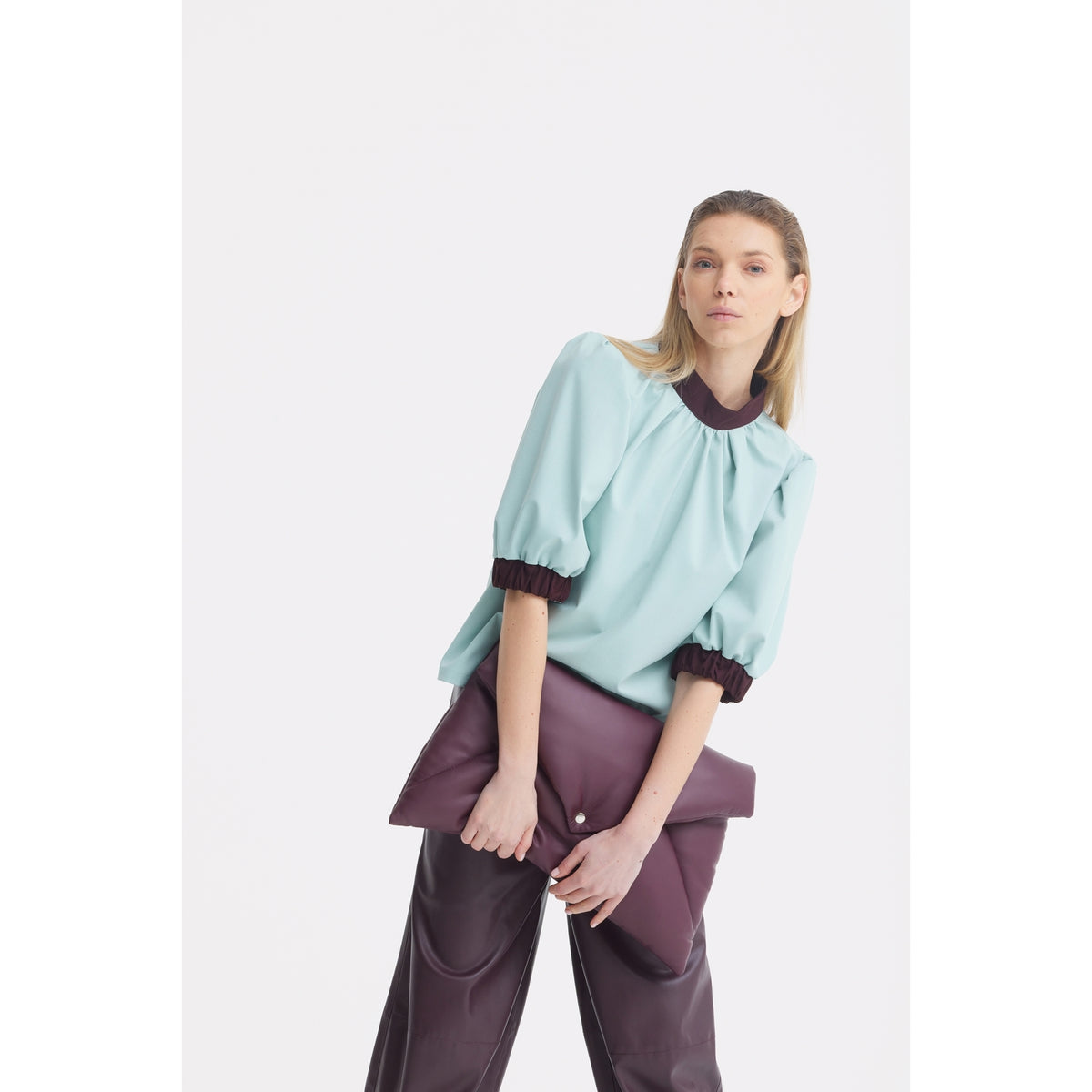 A woman with straight blonde hair wears a JULIA ALLERT Textured Short Sleeve Blouse in light blue with puffed sleeves and dark brown cuffs, paired with brown pants. She holds a large purple clutch and stands against a plain white background.