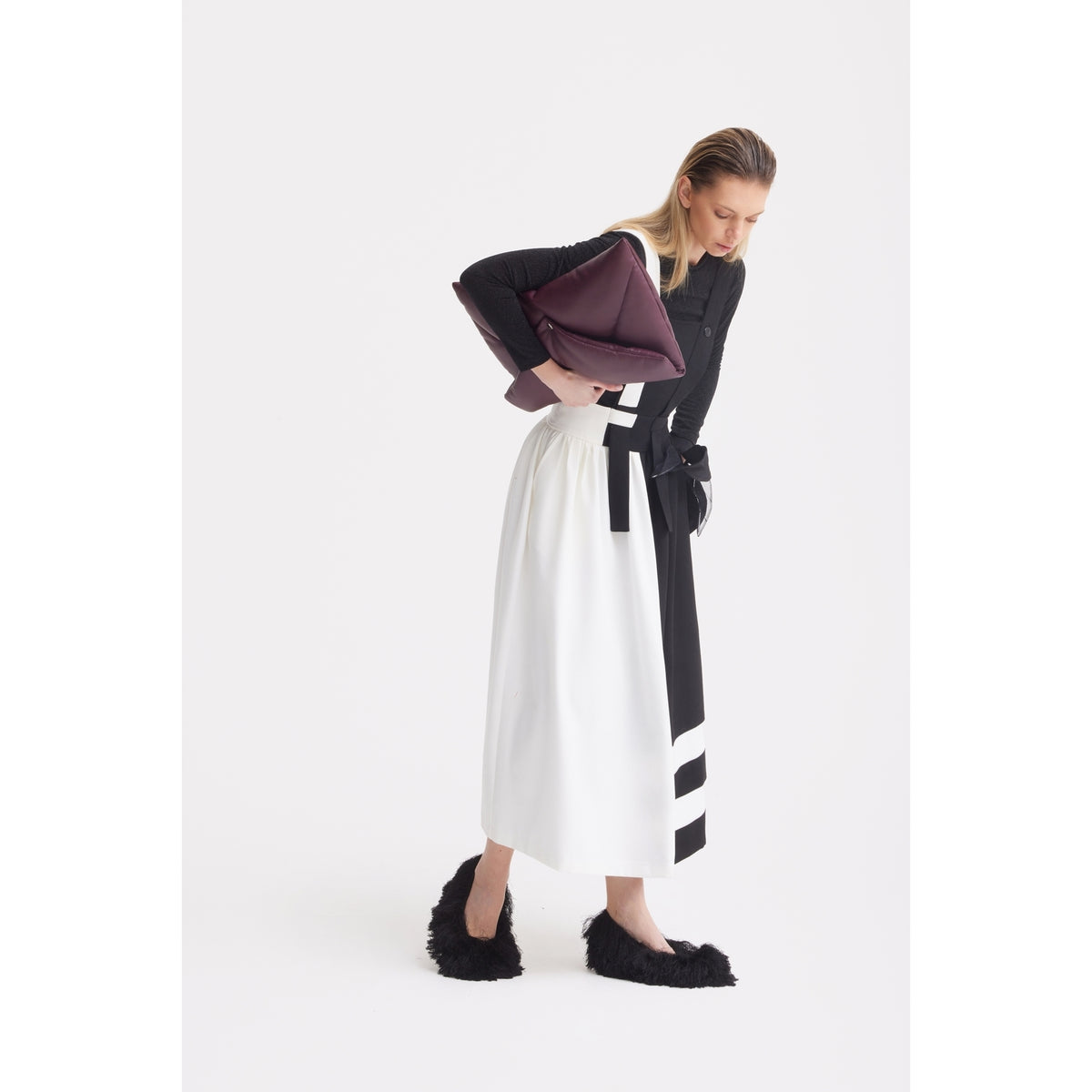 A woman wears the JULIA ALLERT Midi Apron Dress With Adjustable Straps in Black Ivory, paired with a long black-and-white skirt, a large burgundy clutch, and black fuzzy slippers, standing against a plain white background.